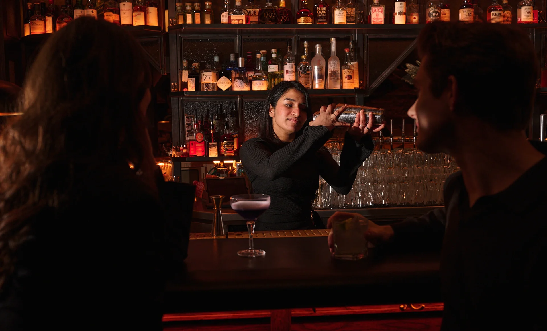 Bartender shaking a cocktail shaker behind a bar with two customers holding drinks.