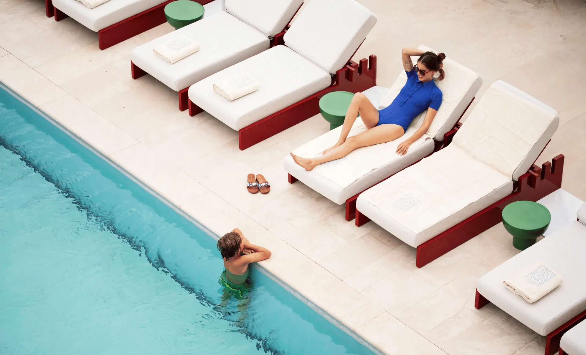 Woman in blue swimsuit on lounge chair talking to boy in green swim trunks in pool.