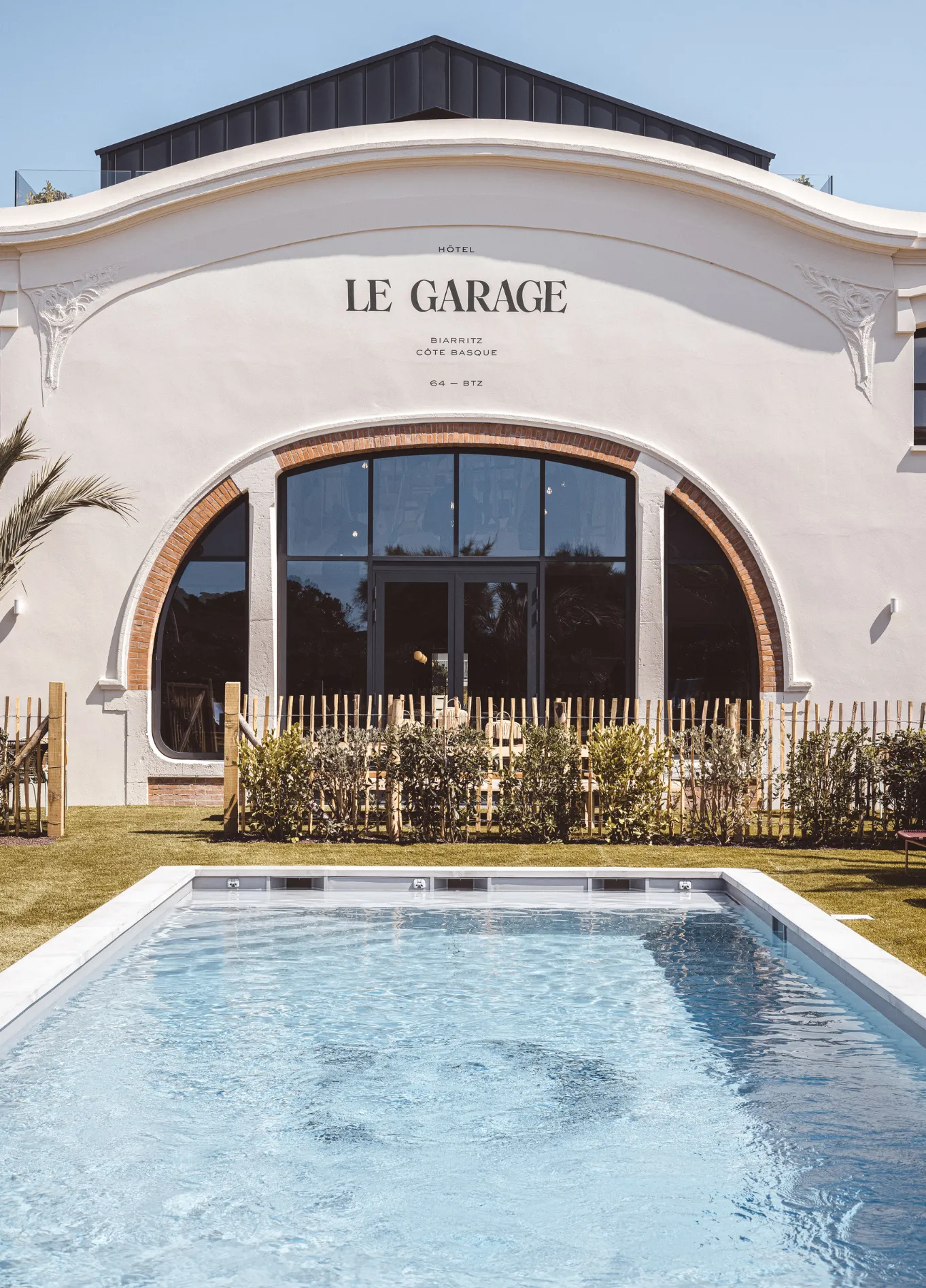 Facade of "Le Garage" building featuring a large arched window and a swimming pool in the foreground.