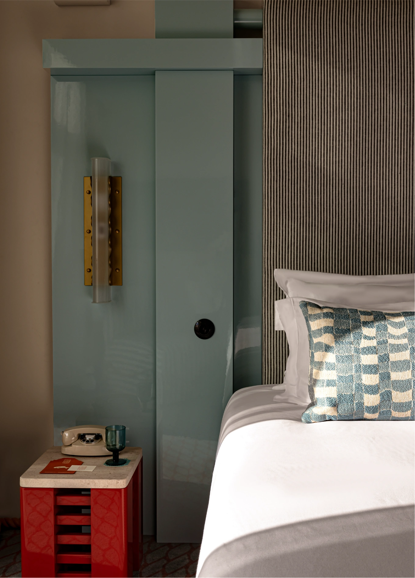 Detail of a modern hotel room featuring a red bedside table, a comfortable bed, and a blue-gray sliding panel on the wall.