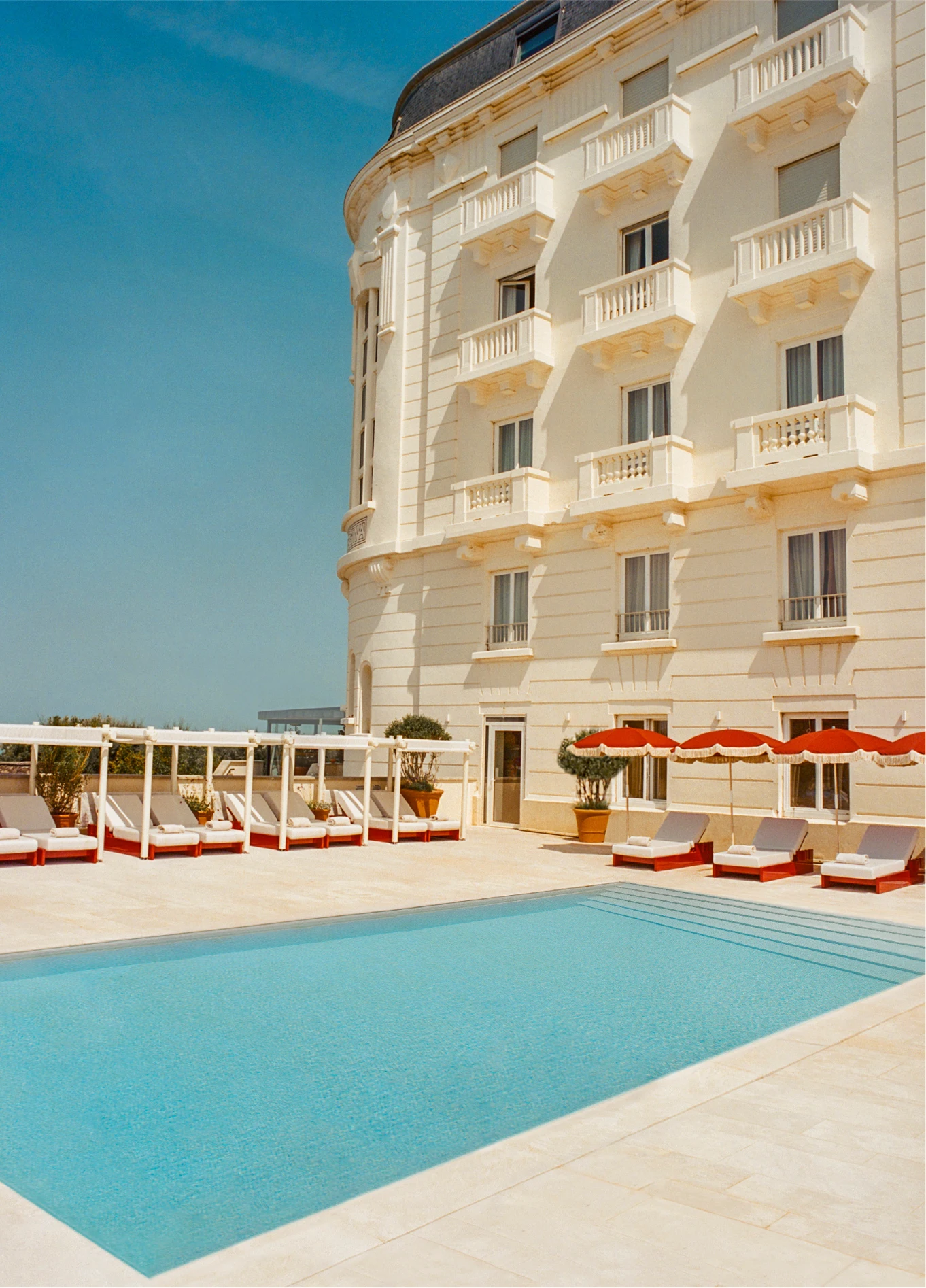 Rectangular swimming pool surrounded by a terrace, with lounge chairs under red umbrellas in front of an imposing and bright hotel facade.