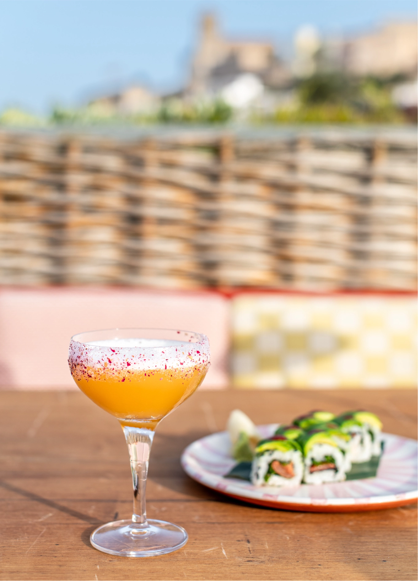An orange cocktail in a coupe glass and a plate of sushi placed on an outdoor table, with a terrace setting in the background.