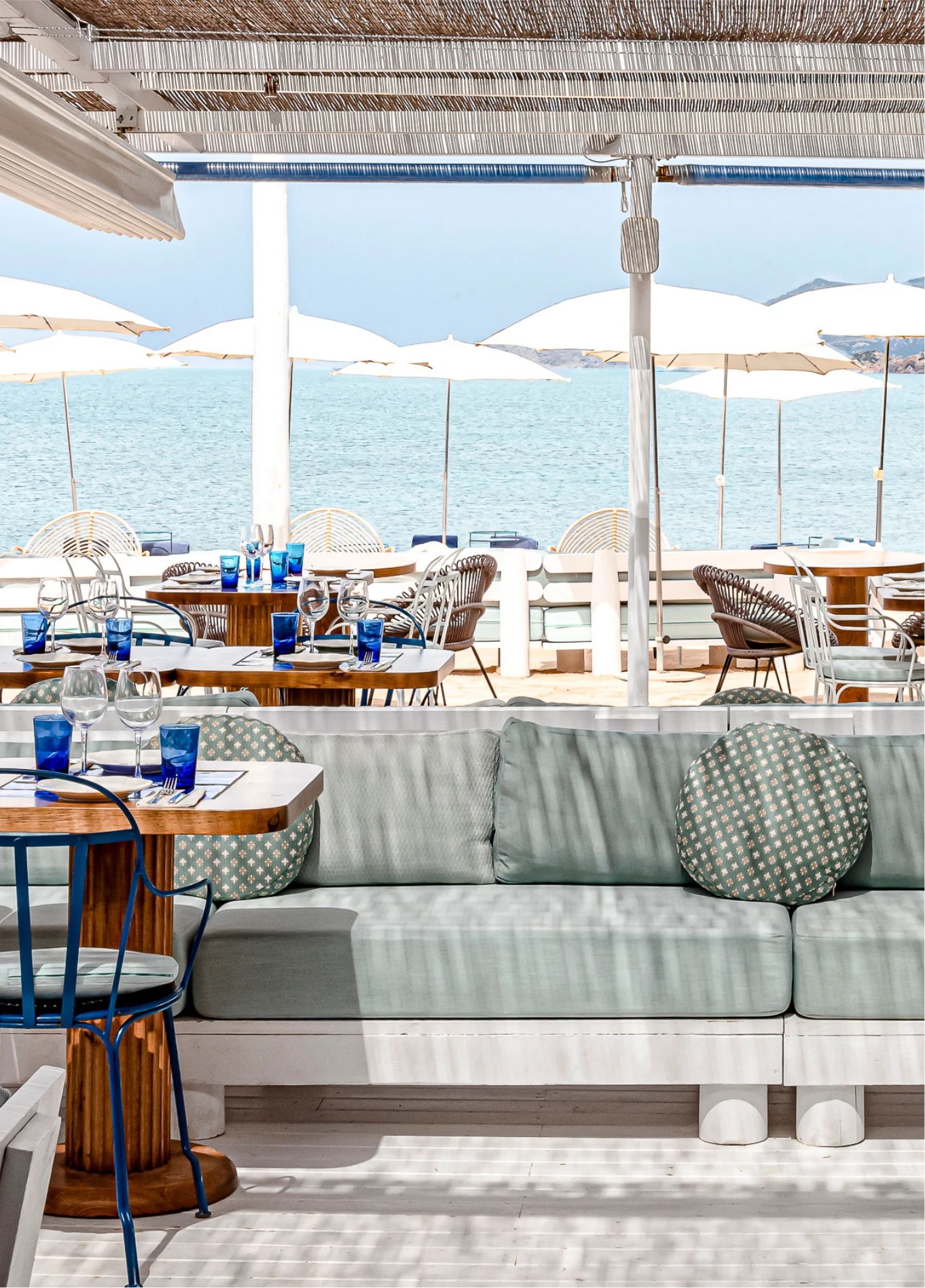 A restaurant terrace with comfortable sofas and tables, offering a view of the sea with many white umbrellas.