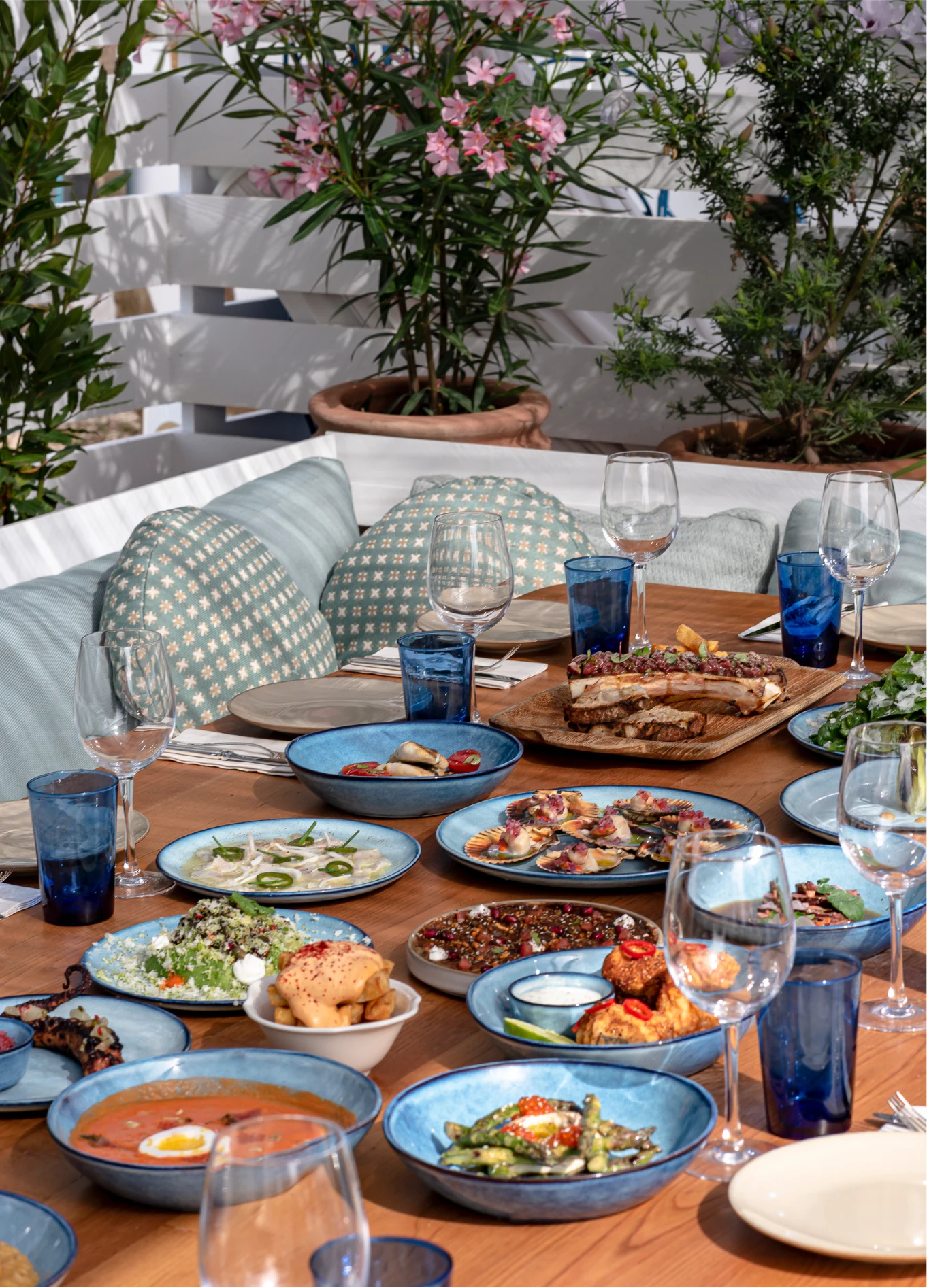 A wooden table set for a meal, filled with many varied dishes and stemmed glasses, in an outdoor setting.