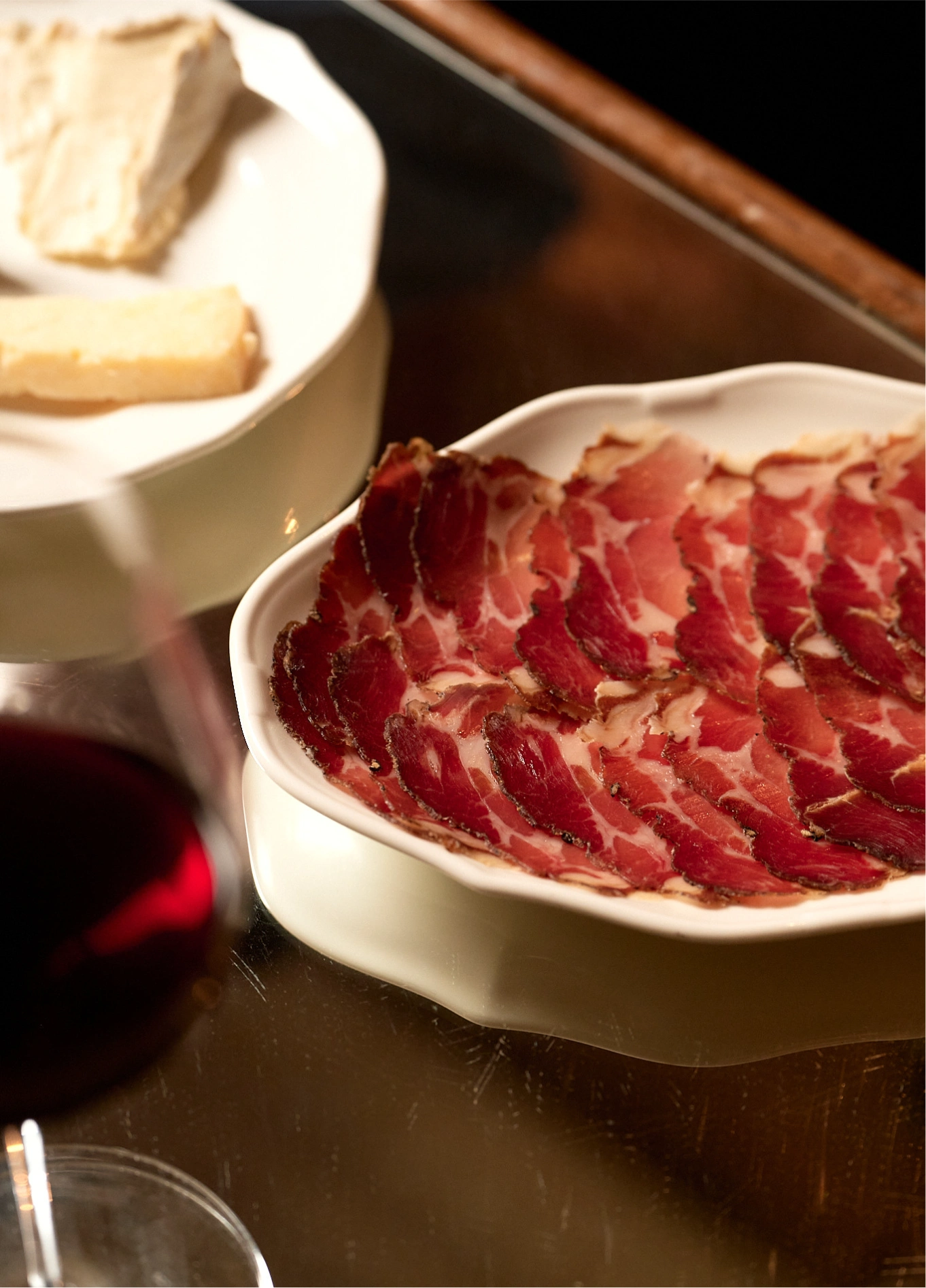 Plate of thinly sliced charcuterie presented alongside a cheese platter and a glass of red wine.