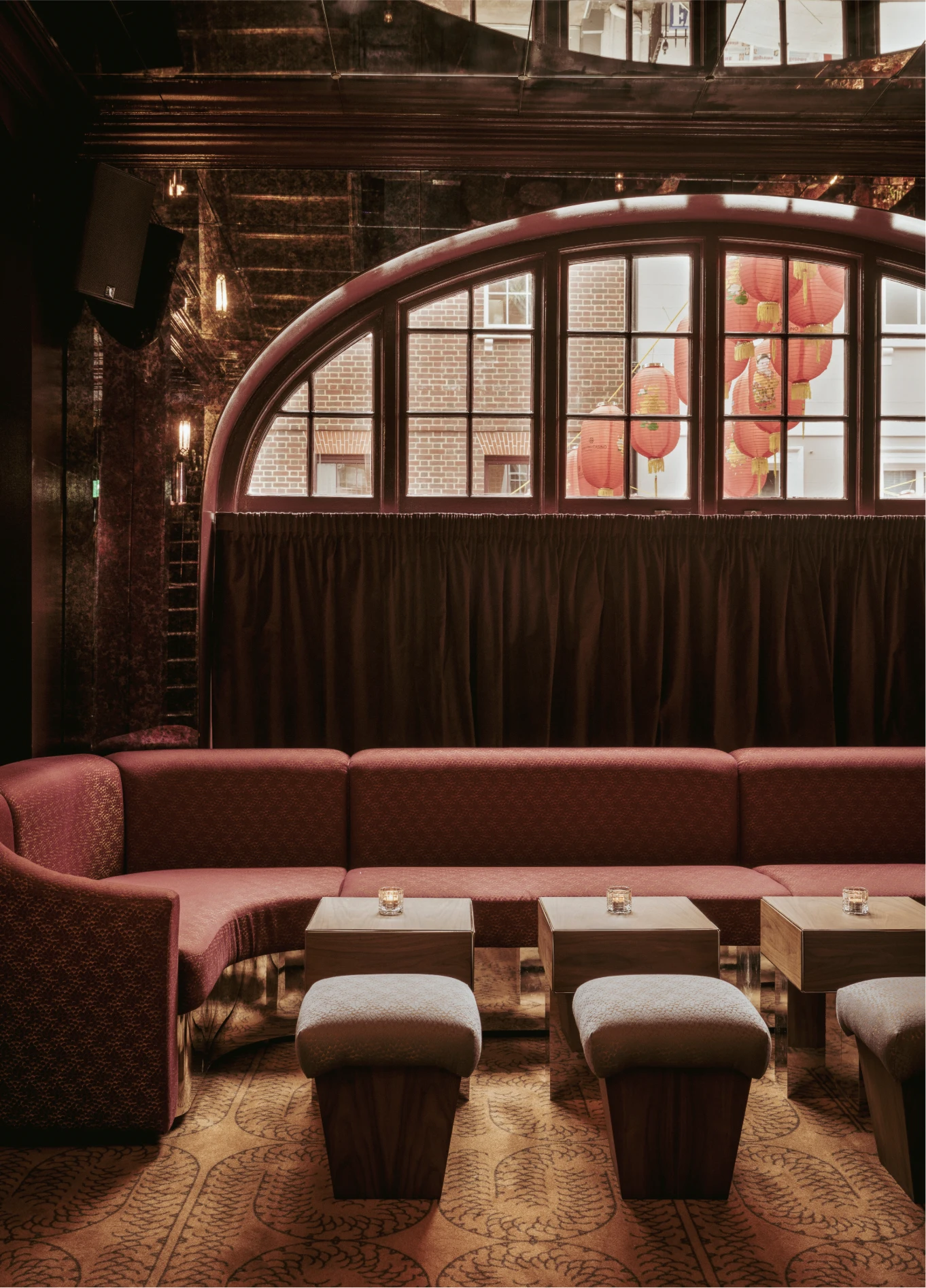 A cozy lounge area with large red sofas and coffee tables in front of a large arched window at the Experimental Cocktail Club.