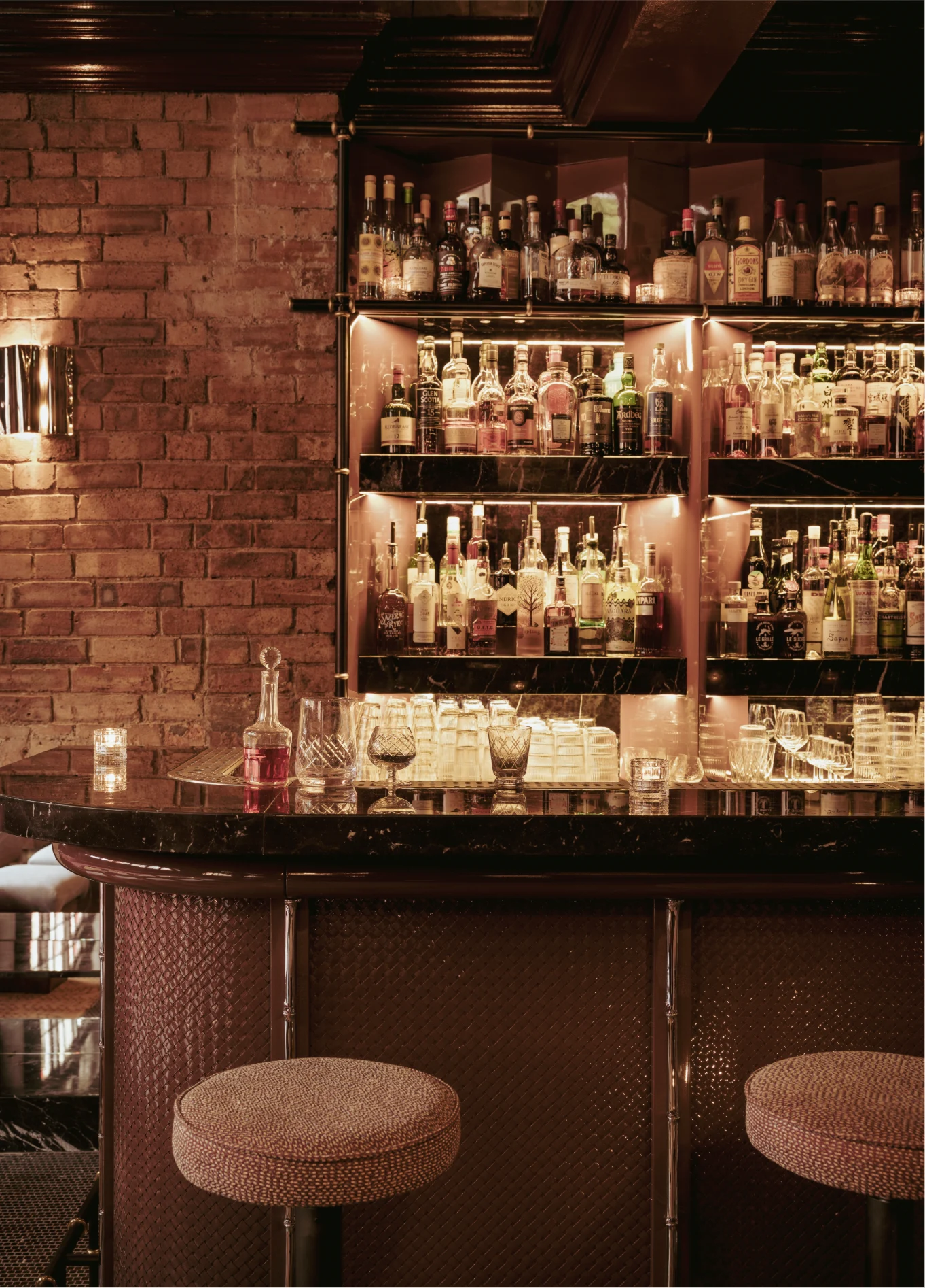 An elegant bar counter with stools and shelves filled with spirit bottles, in front of a brick wall at the Experimental Cocktail Club.