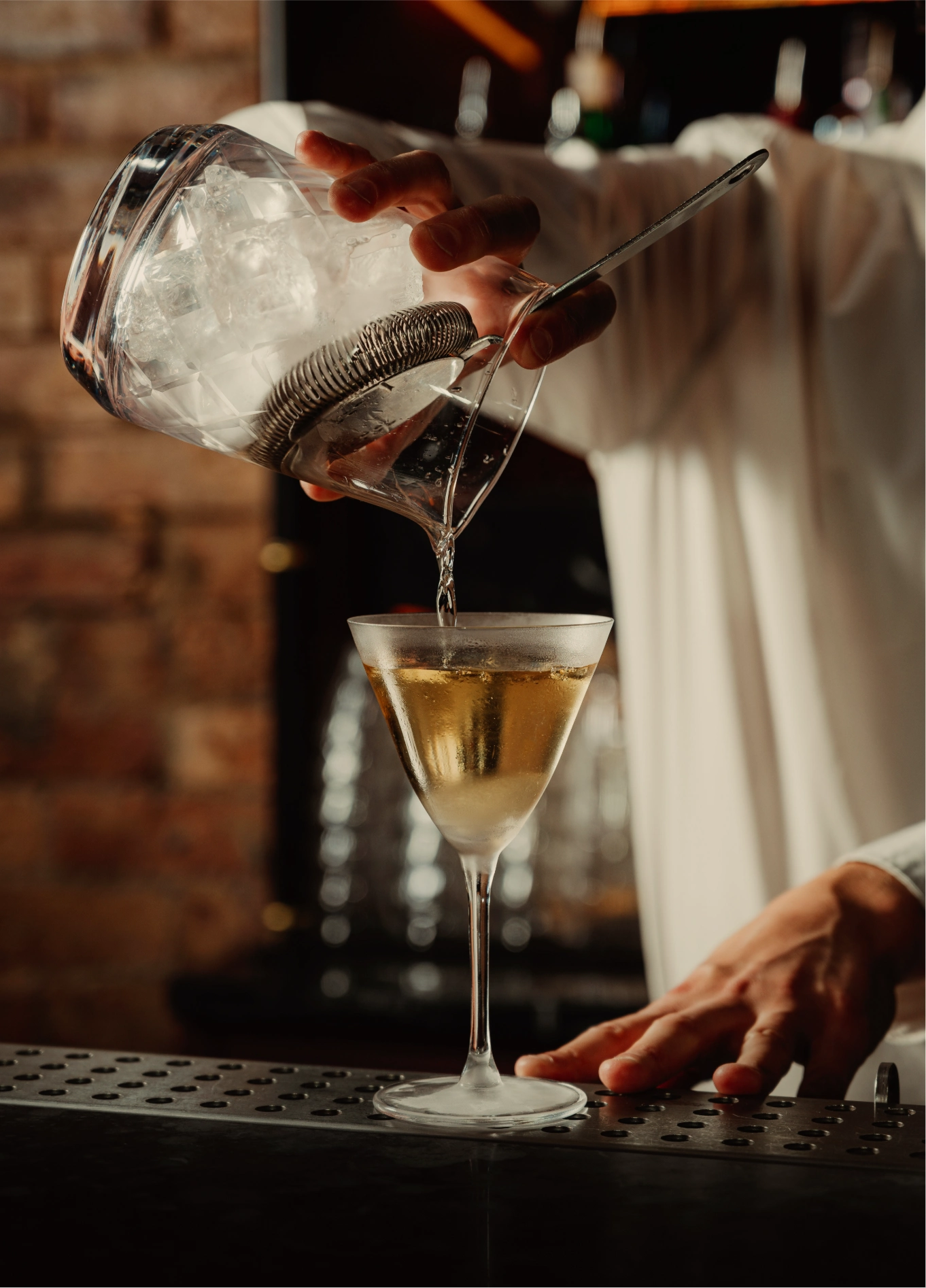 A bartender pouring a cocktail into a stemmed glass from a shaker, with an exposed brick wall in the background at the Experimental Cocktail Club.