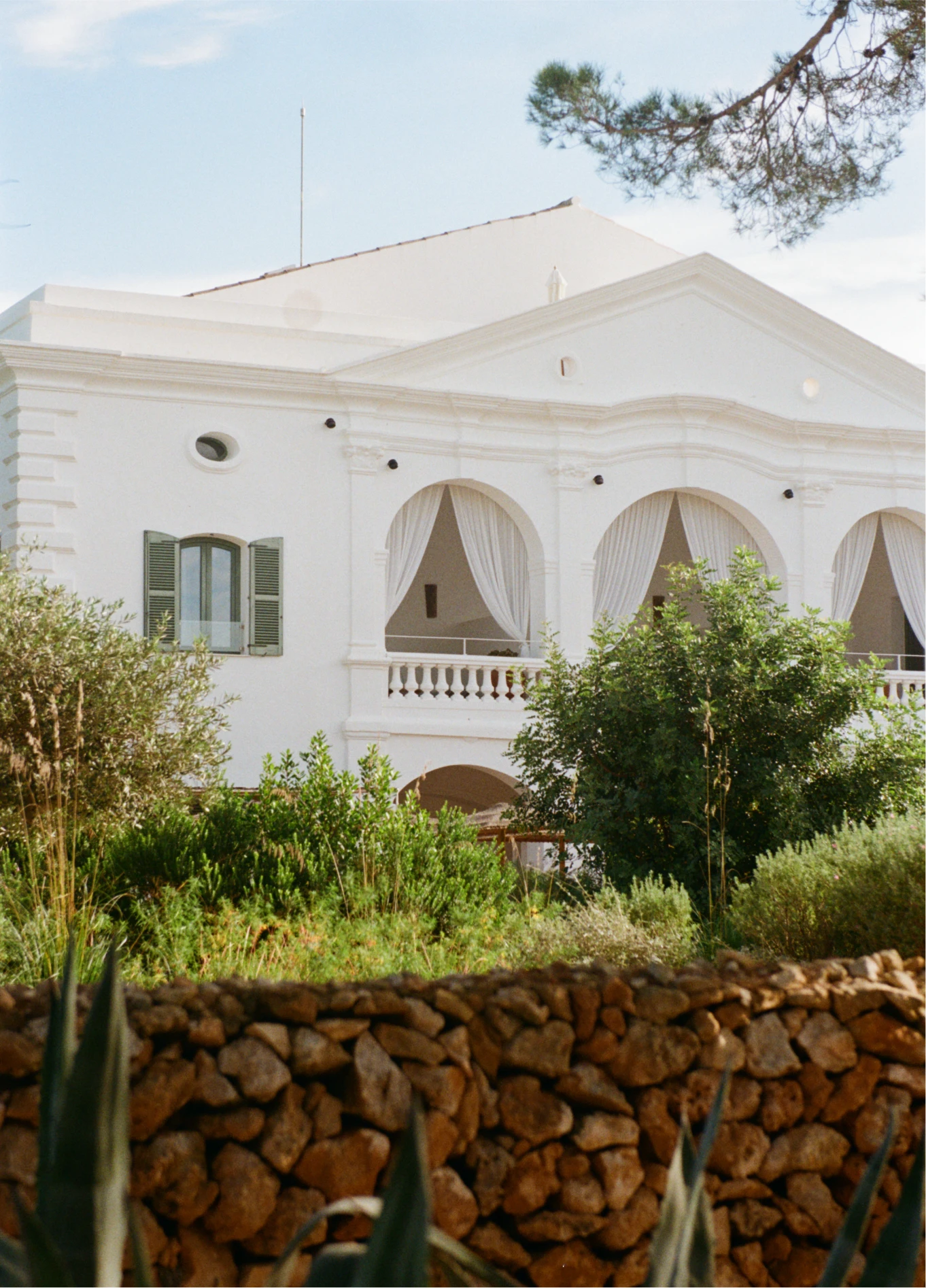 White facade of Menorca Experimental, characterized by its architectural arches, surrounded by Mediterranean vegetation and a dry stone wall in the foreground.