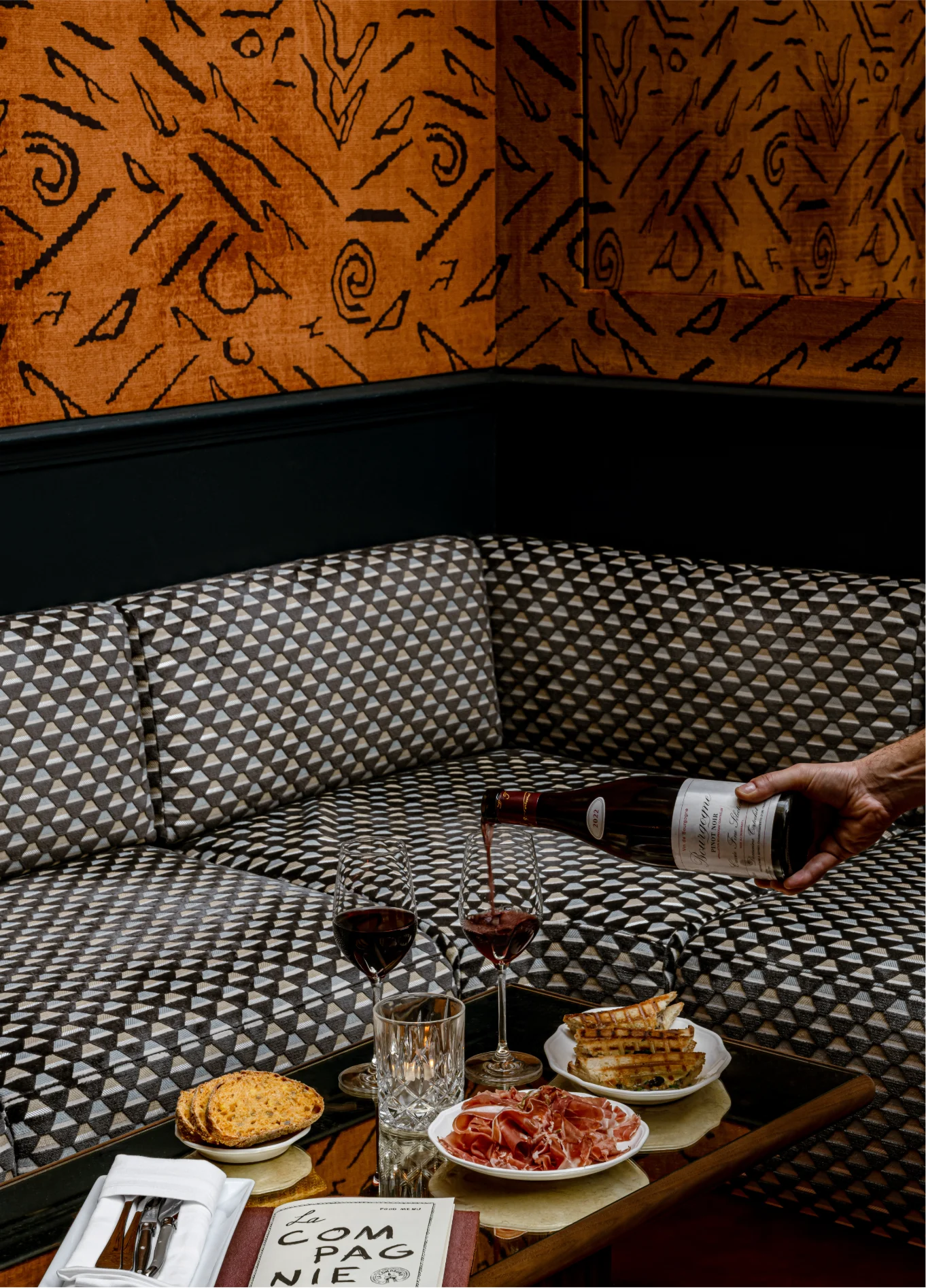 A hand pouring wine into a glass on a coffee table, in front of a checkered sofa, in a space with a wall decorated with orange graphic patterned wallpaper.