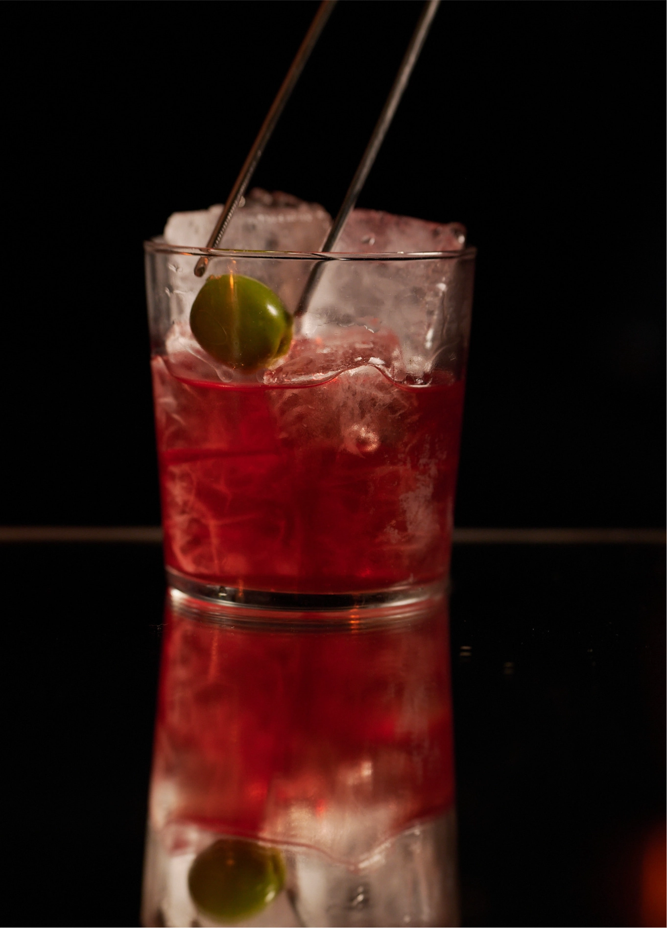 A hand using tongs to add an olive to an iced red cocktail at the Experimental Cocktail Club.