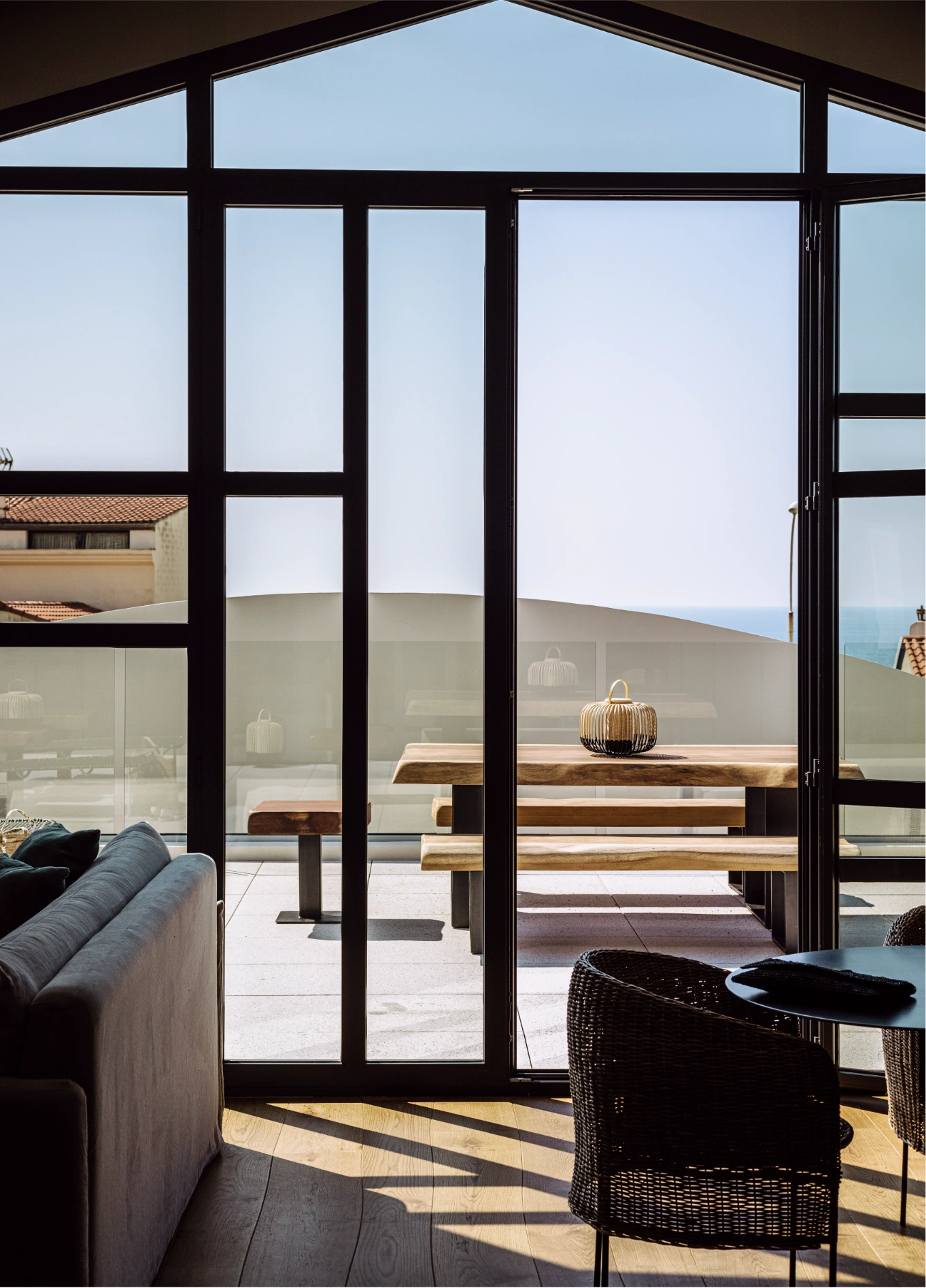 Interior view through a large glass window looking out onto a terrace with a wooden table and an open landscape.