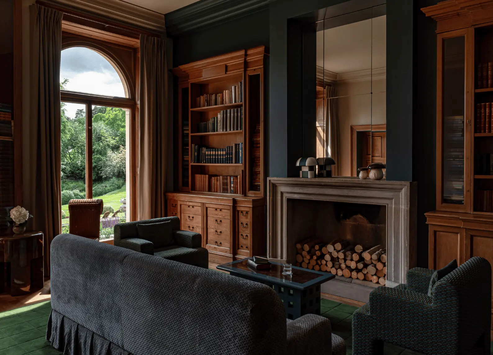 Wooden library with fireplace, grey armchairs and arched window overlooking green park at Cowley Manor Experimental, Cotswolds