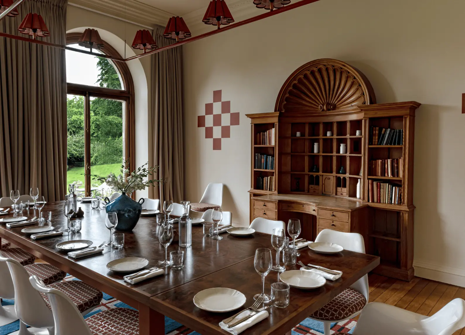 Private dining room with large wooden table, white chairs and arched window overlooking the garden at The Study, Cowley Manor Experimental, Cotswolds