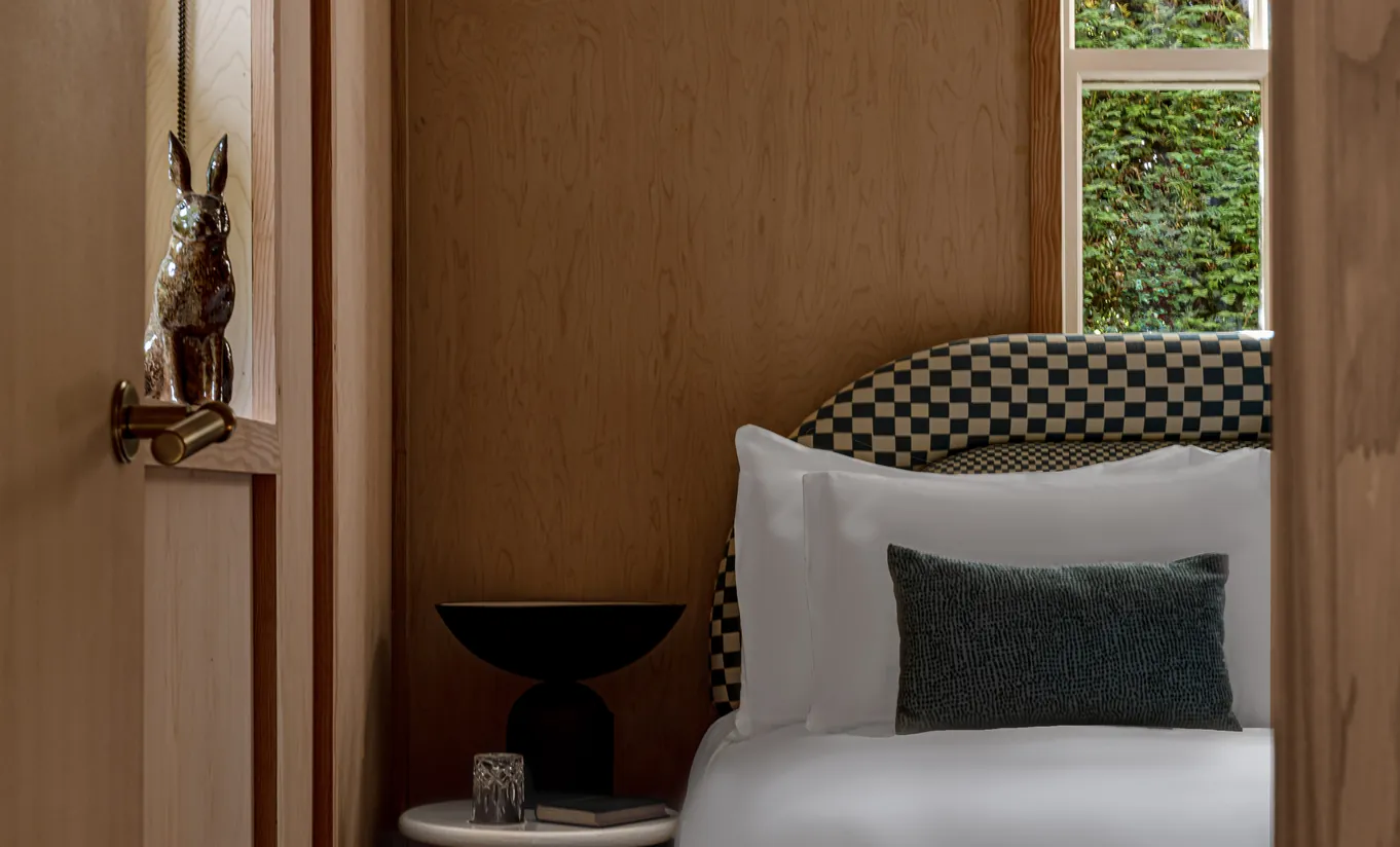 Detail of a bed with white linen, patterned cushions and a wooden sculpture in the Grand Cowley Manor Suite at Cowley Manor Experimental, Cotswolds