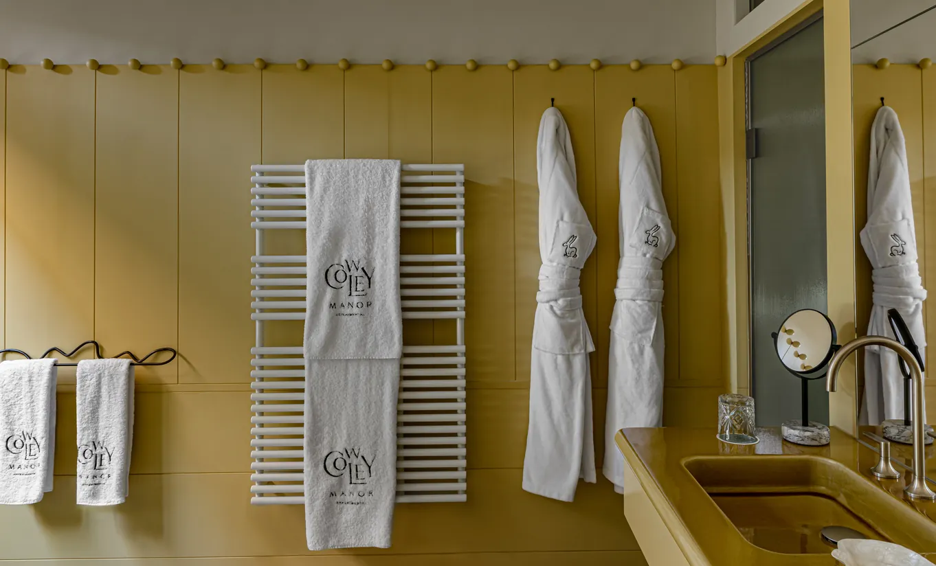Bathroom with mustard yellow walls, white monogrammed bathrobes and a heated towel rail in the Treehouse room at Cowley Manor Experimental, Cotswolds