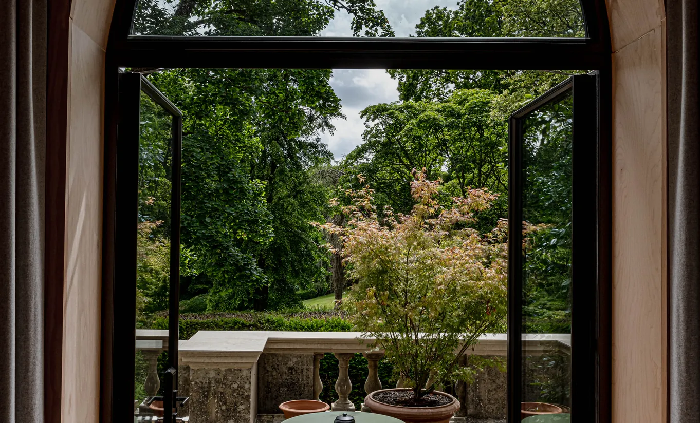 Arched window opening onto a lush private terrace surrounded by trees in the Waterlily with Terrace room at Cowley Manor Experimental, ideal for a countryside getaway in the Cotswolds