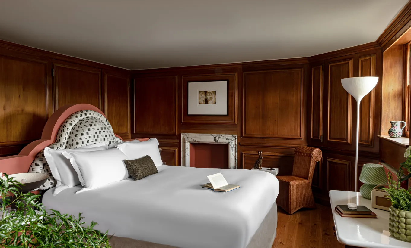 Bedroom with a white double bed, rich dark wood panelling, a fireplace and lush indoor plants in the Water Lily room at Cowley Manor Experimental, Cotswolds