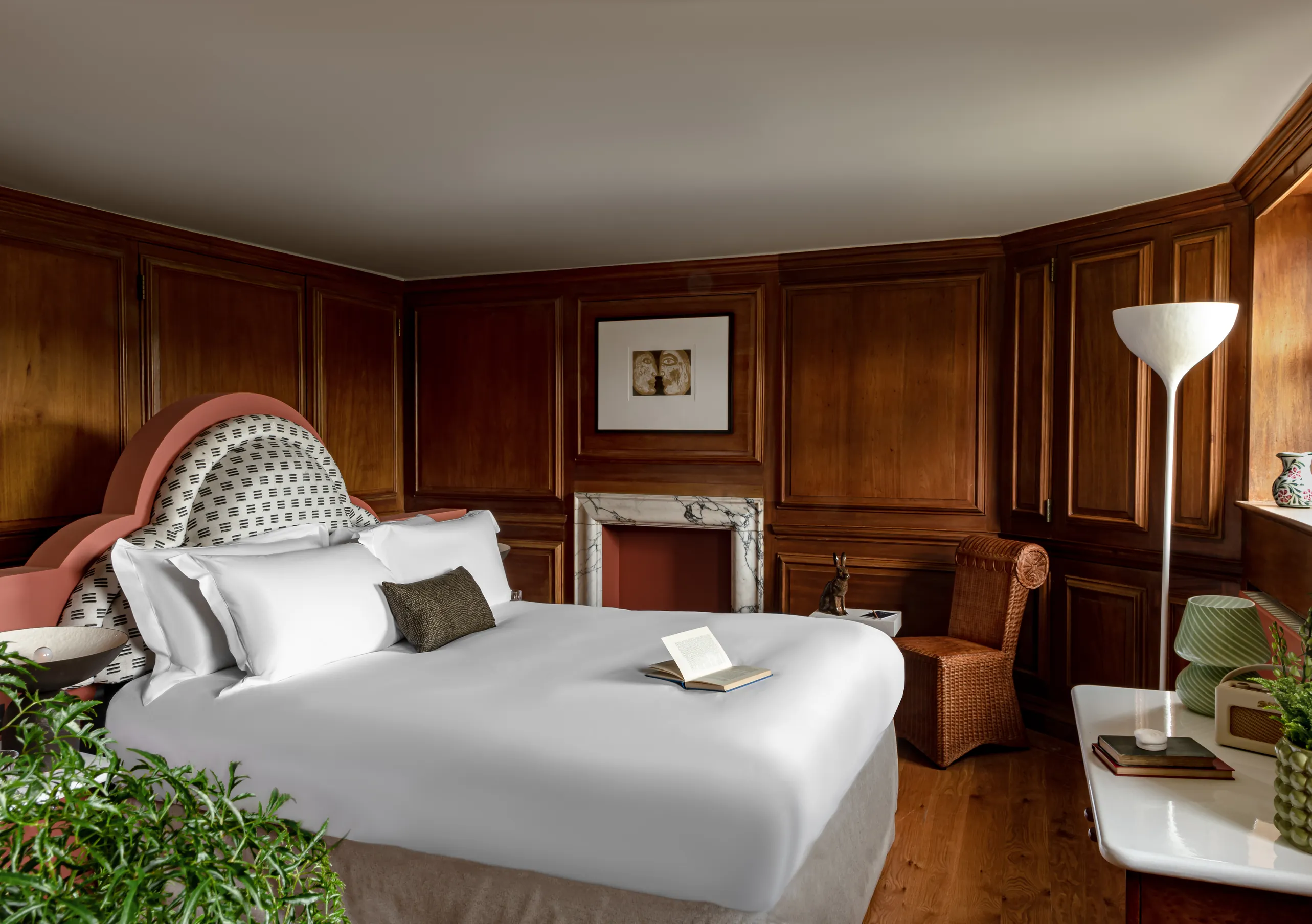 Bedroom with a white double bed, rich dark wood panelling, a fireplace and lush indoor plants in the Water Lily room at Cowley Manor Experimental, Cotswolds
