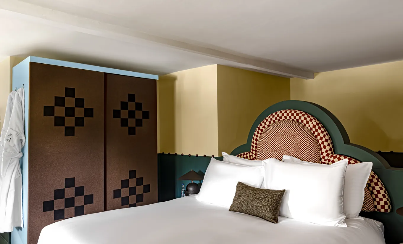 Bedroom with a white double bed, a mosaic headboard, mustard yellow walls and a checkered wardrobe in the Wildflower room at Cowley Manor Experimental, Cotswolds