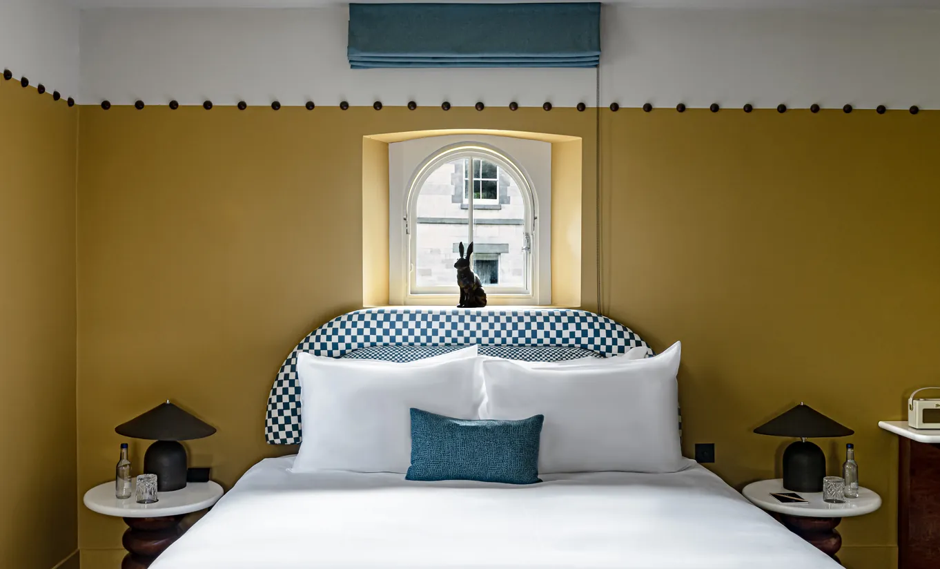 Bright bedroom with a white double bed, mustard yellow walls and symmetrical bedside lamps in the Wildflower room at Cowley Manor Experimental, Cotswolds
