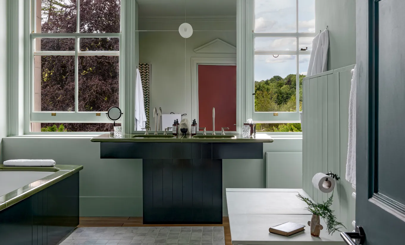 Bright bathroom with sage green cabinetry, a double sink and a window overlooking the Cotswolds countryside in the Swan Lake Suite at Cowley Manor Experimental