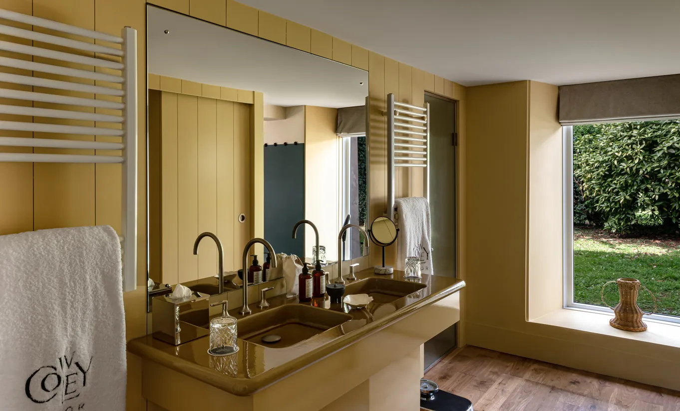 Bathroom with mustard yellow walls, a wooden vanity unit and a window overlooking the garden in the Wildflower with Terrace room at Cowley Manor Experimental, Cotswolds