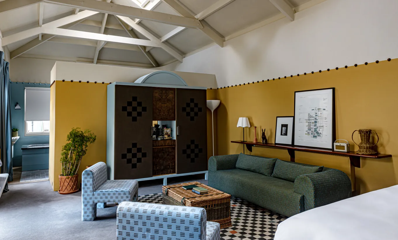 Spacious bedroom with vaulted ceilings, mustard yellow walls, a green sofa and a patterned rug in the Churns River Junior Suite at Cowley Manor Experimental, countryside retreat in the Cotswolds