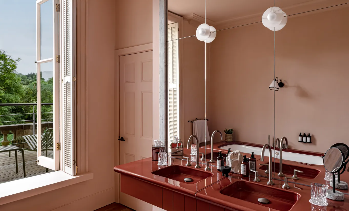 Bright bathroom with terracotta walls, a red double sink and an open window with a garden view in the St Mary Suite at Cowley Manor Experimental, Cotswolds