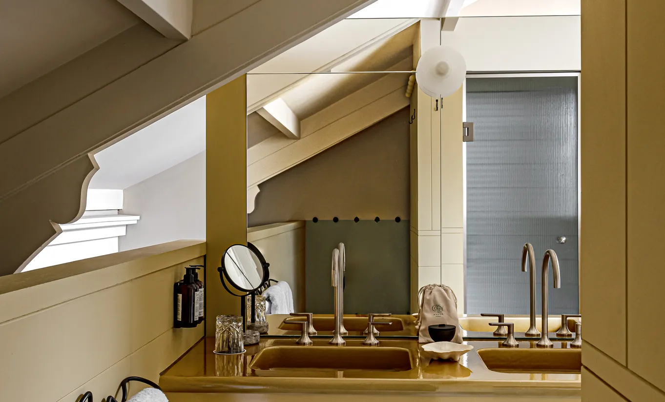 Compact bathroom with a built-in bathtub, wooden shelving and a vanity mirror under a sloped ceiling in the Alpaca's Room at Cowley Manor Experimental, Cotswolds