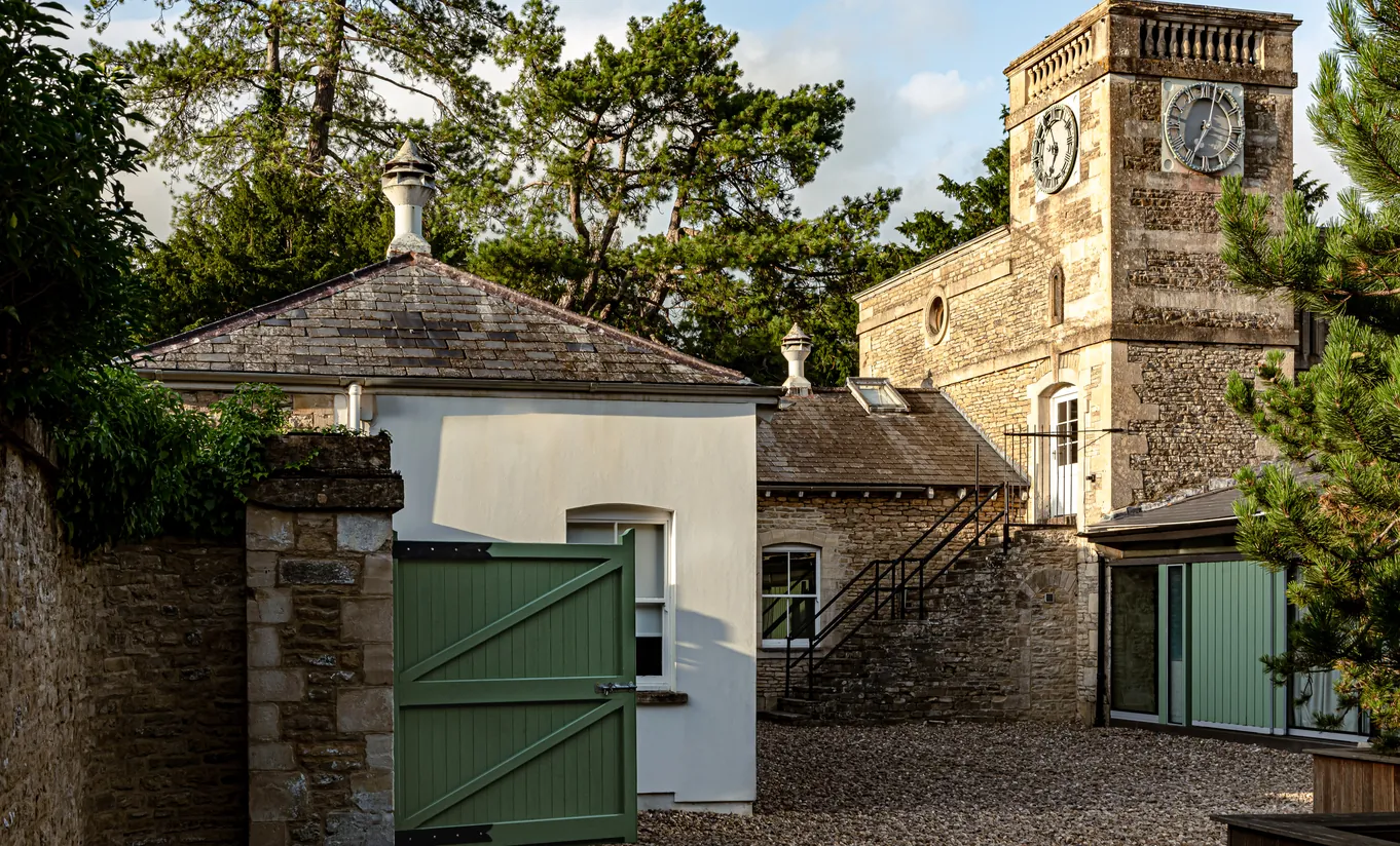Exterior view of the stone clock tower and manor outbuildings with a green gate at Cowley Manor Experimental, a luxury countryside hotel in the Cotswolds