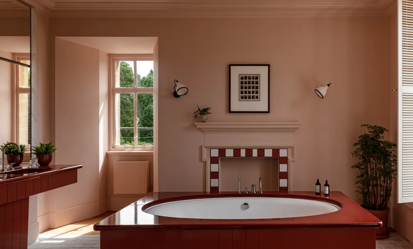 Bathroom with a oval bathtub, terracotta walls and a red and white tiled fireplace in the Cowley Manor Suite at Cowley Manor Experimental, Cotswolds