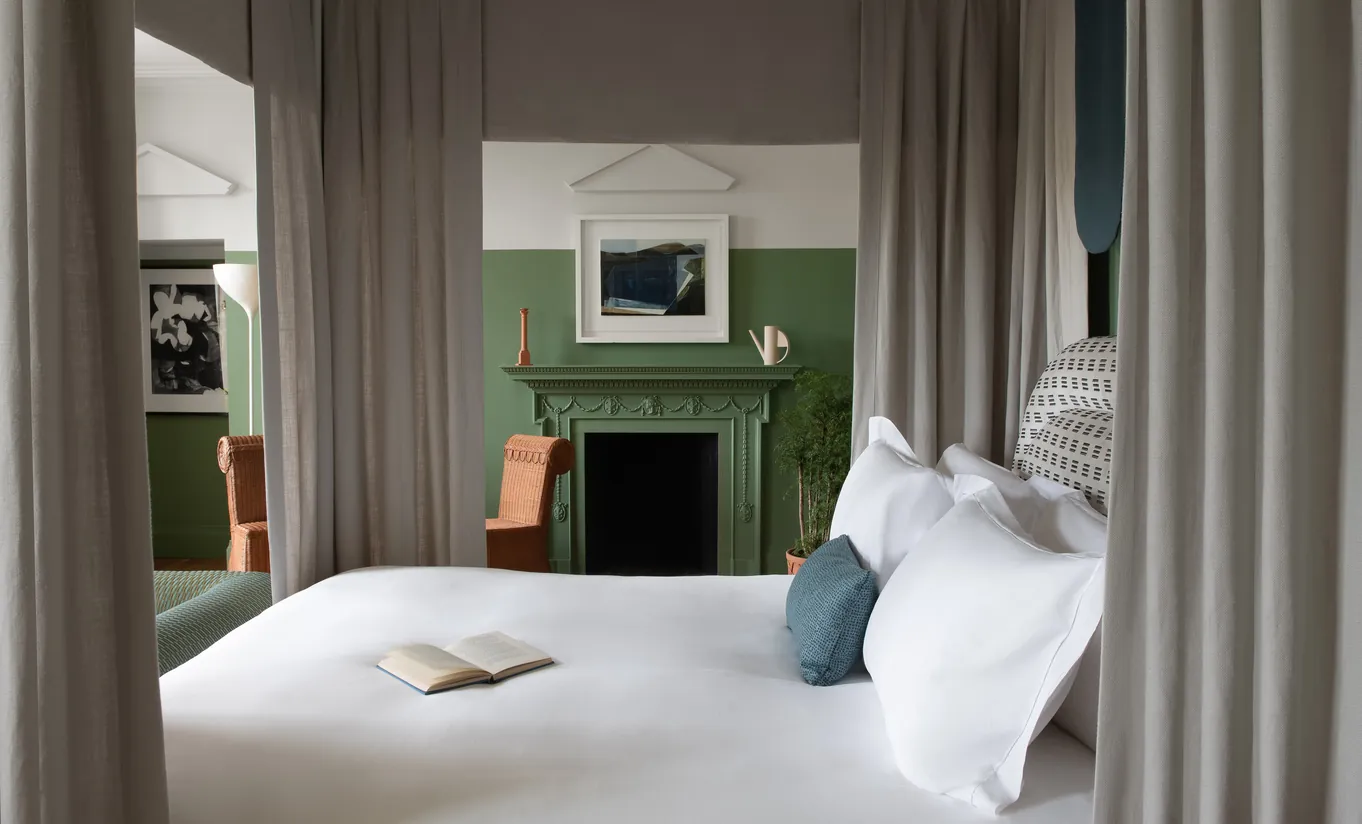 Bedroom with a white double bed, green walls, a fireplace and floor-length curtains in the Cowley Manor Suite at Cowley Manor Experimental, Cotswolds countryside retreat