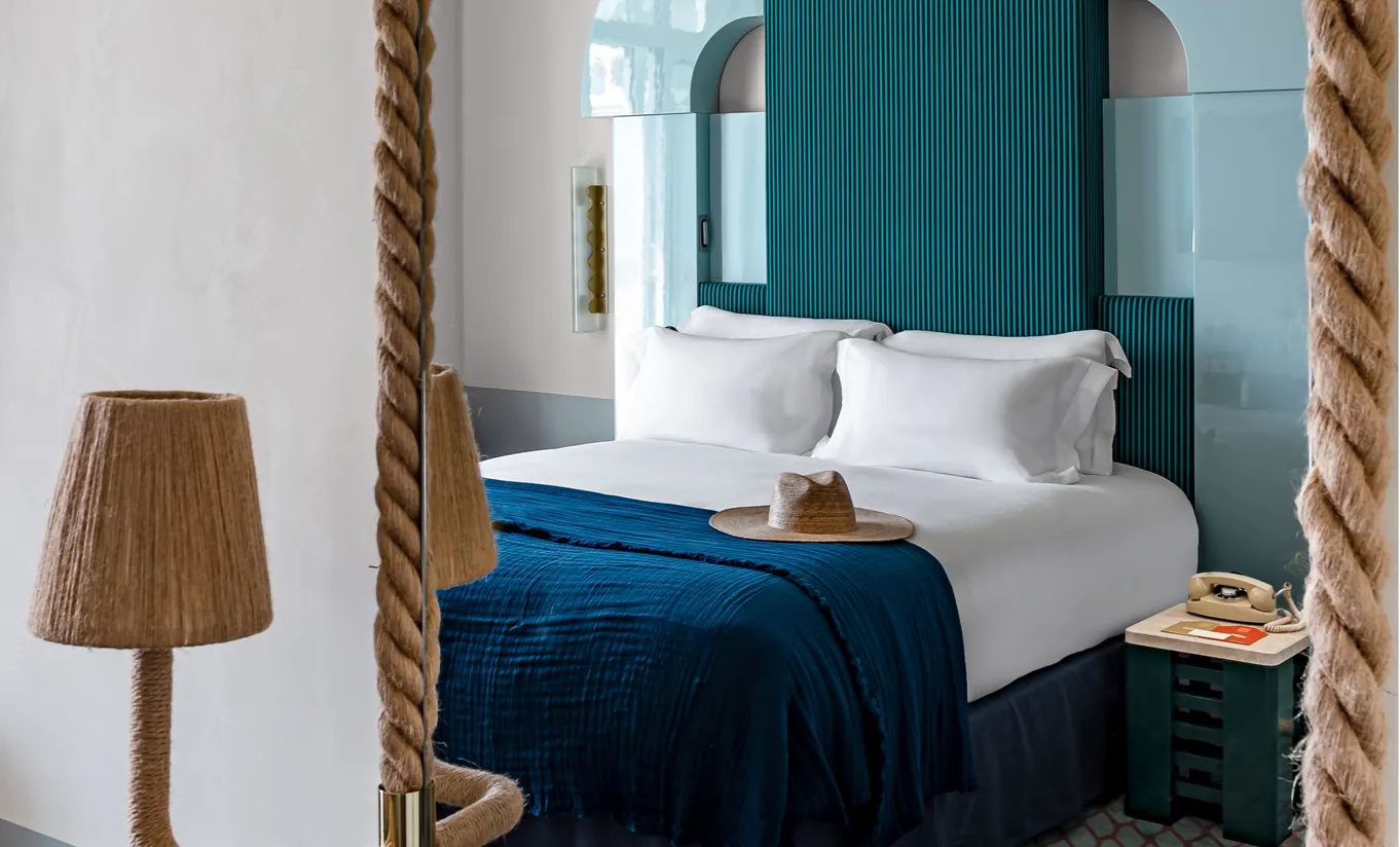 Experimental Group hotel bedroom with teal green headboard, white bedding, blue throw blanket, and rope bed post detail