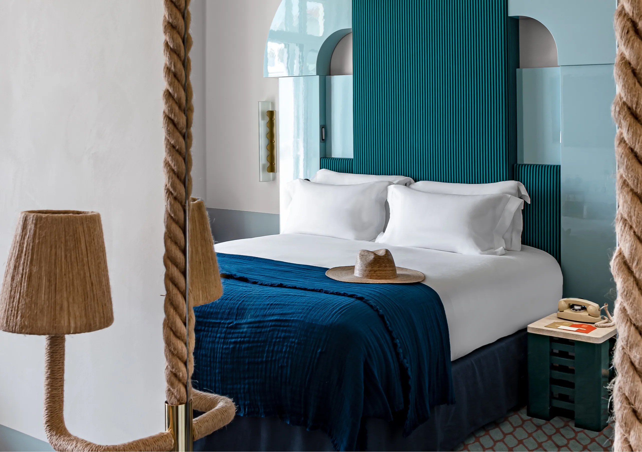 Experimental Group hotel canopy bed with teal statement headboard, rope four-poster frame, blue velvet blanket, and warm lamp lighting.
