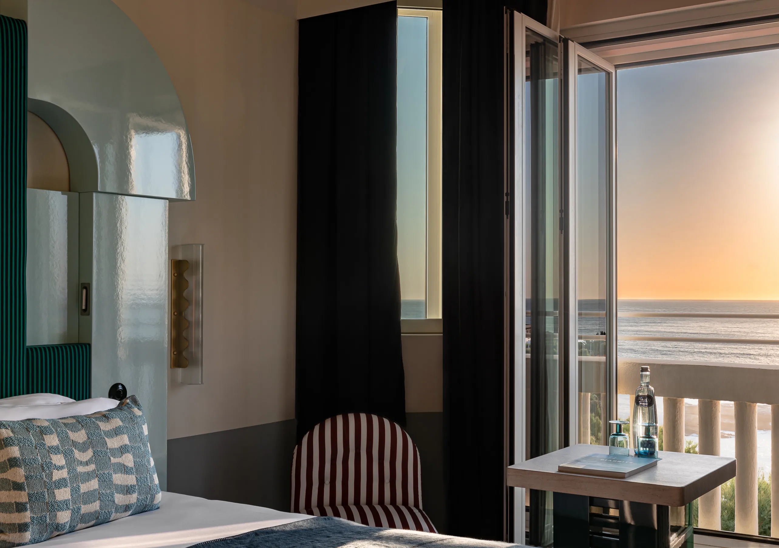 Regina Experimental hotel room at sunset with dark wood wardrobe, striped chair, and dramatic orange ocean view through balcony window.