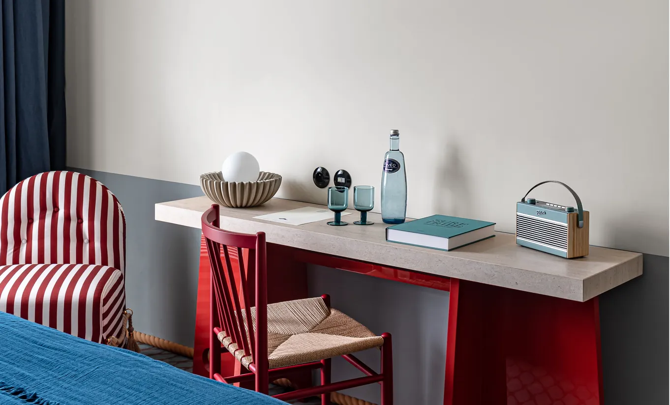 Experimental Paris hotel room desk with red base, concrete surface, woven chair, decorative objects, and blue accent book