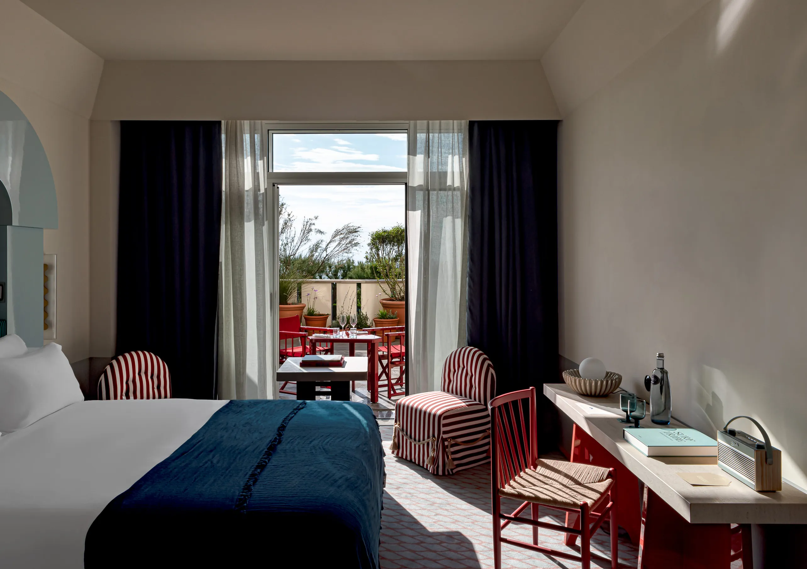 Experimental Paris hotel room with blue bed, navy curtains, red bistro chairs, and open glass doors to a sunlit private balcony.