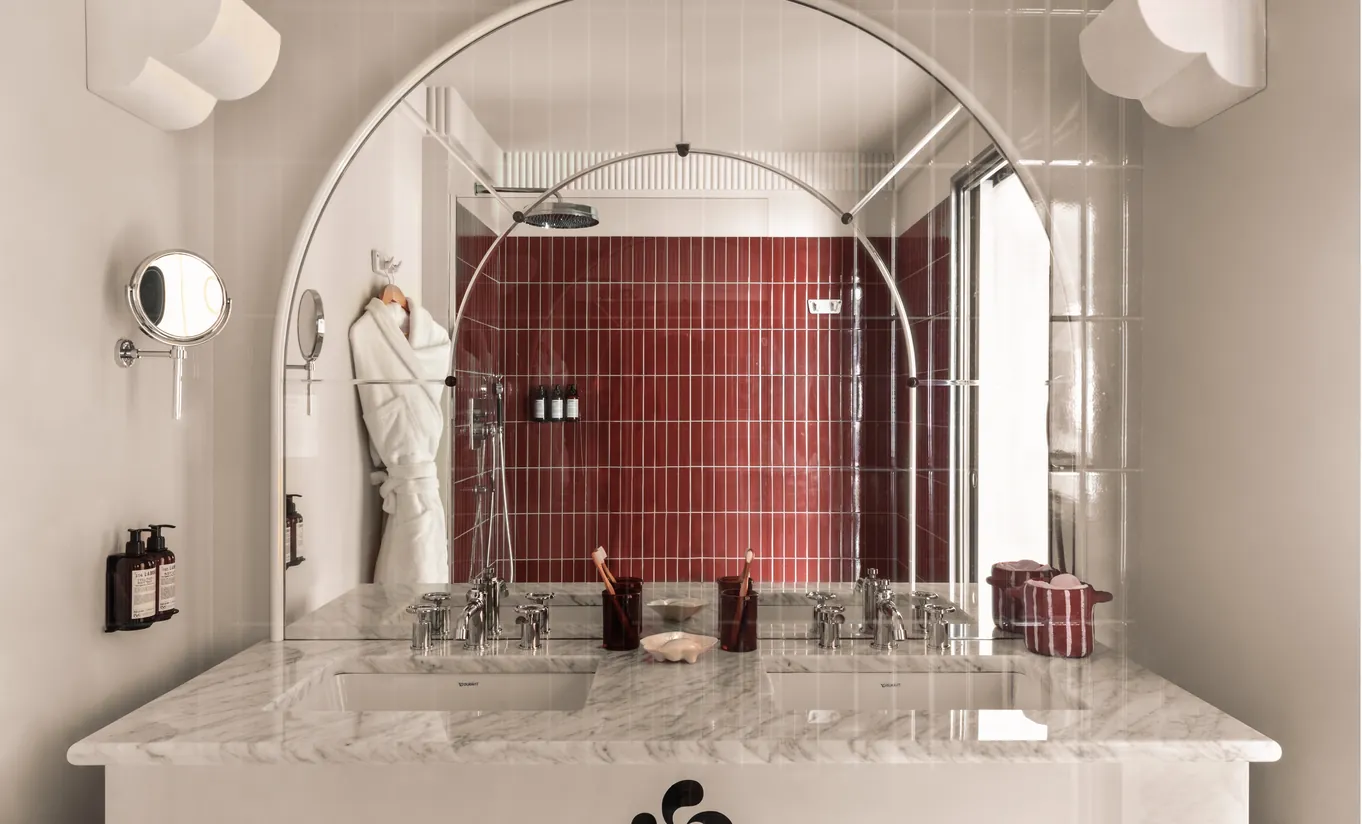 Regina Experimental hotel bathroom with burgundy red mosaic tiled shower, arched mirror, marble sink, and elegant vintage fixtures