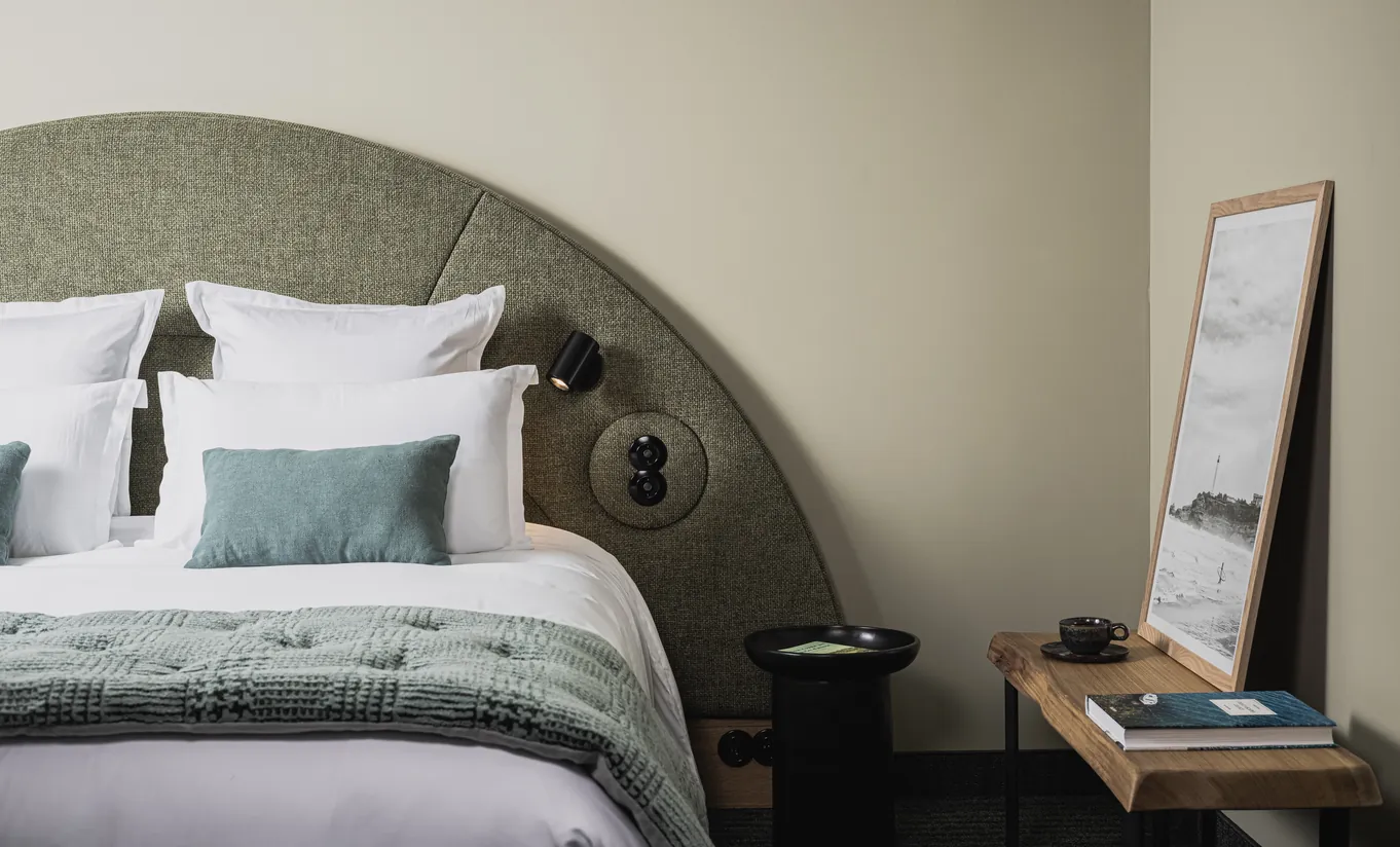Le Garage Biarritz Experimental hotel bedroom with large rounded sage green headboard, white bedding, teal cushion, and minimalist wall art
