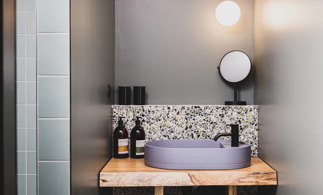 Le Garage Biarritz Experimental hotel bathroom with round grey vessel sink, wood vanity shelf, floral mosaic splashback, and circular mirror