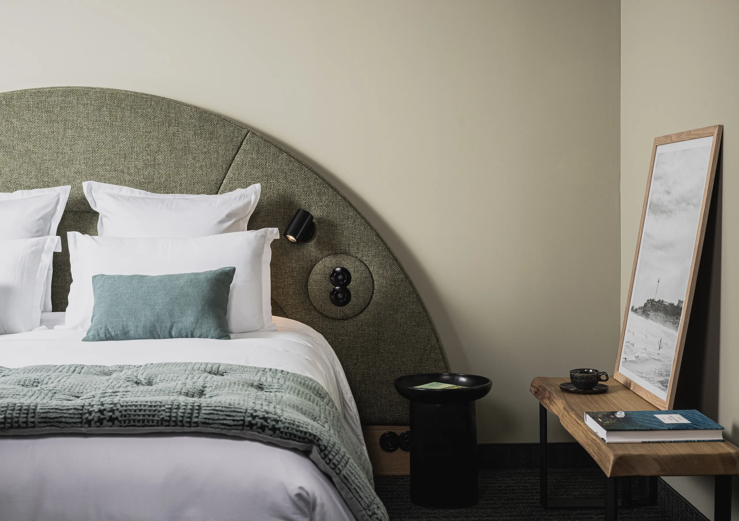 Le Garage Biarritz Experimental hotel bedroom with large rounded sage green headboard, white bedding, teal cushion, and minimalist wall art.
