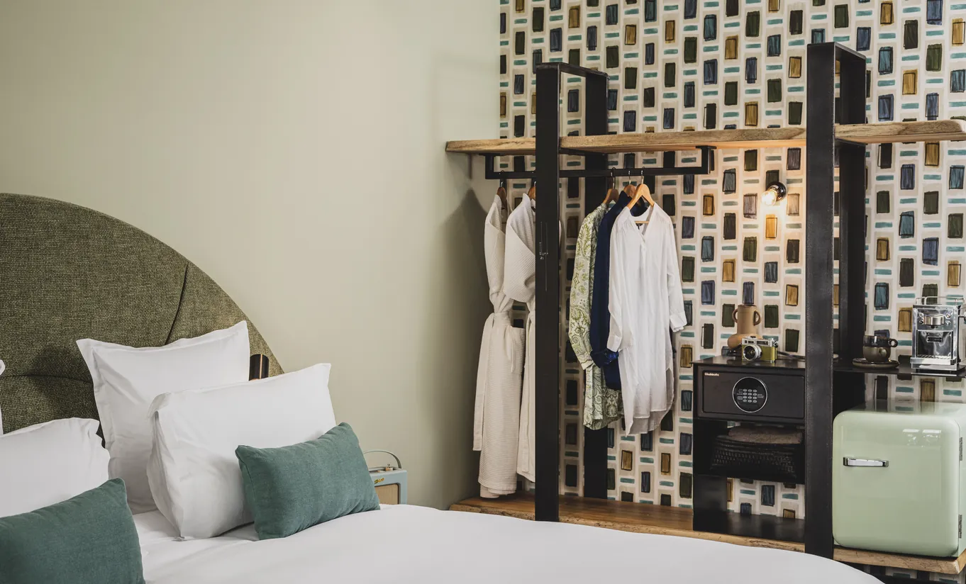 Le Garage Biarritz Experimental hotel room with green headboard, white bedding, open clothing rack with bathrobe, and geometric patterned wallpaper