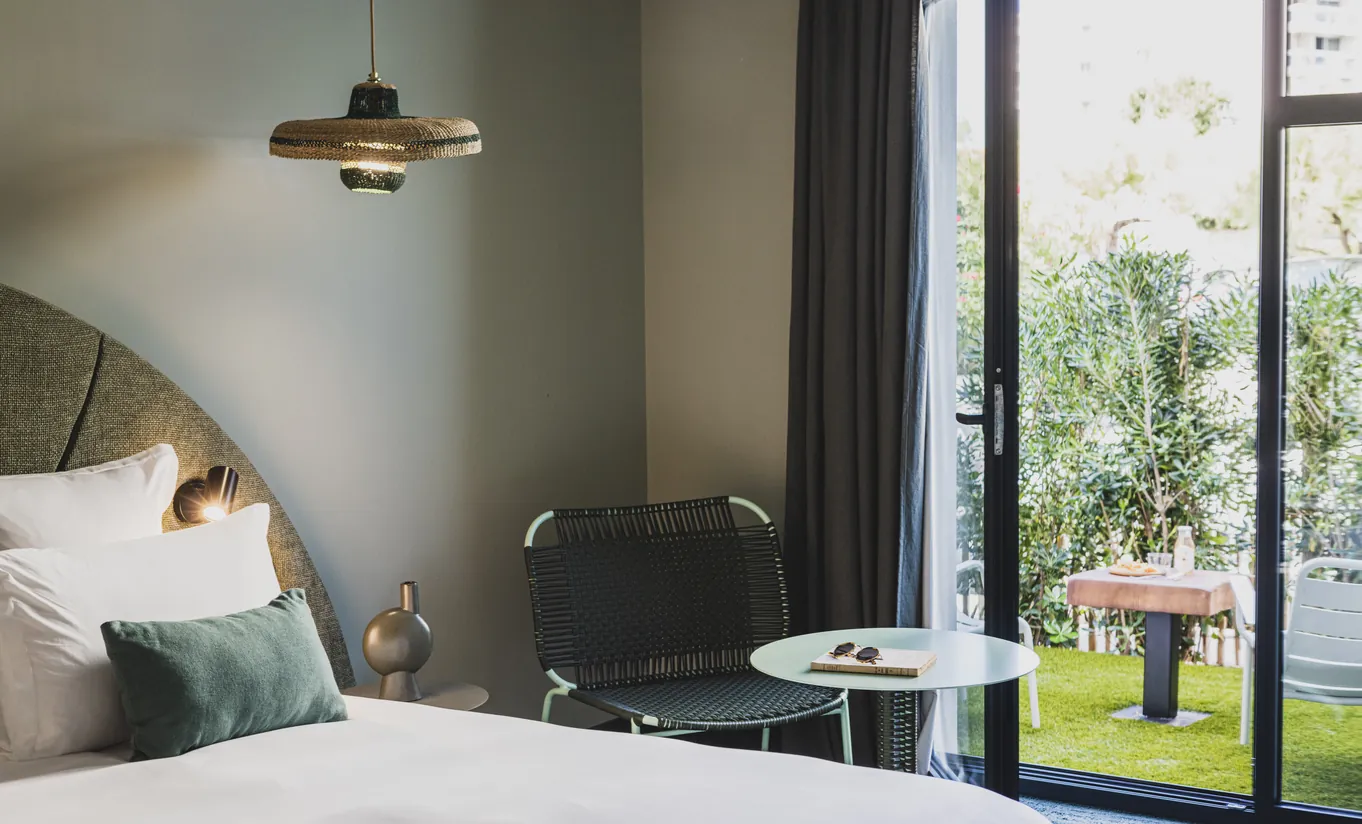 Le Garage Biarritz Experimental hotel room with olive green headboard, white bedding, industrial pendant lamp, and open glass door to private garden