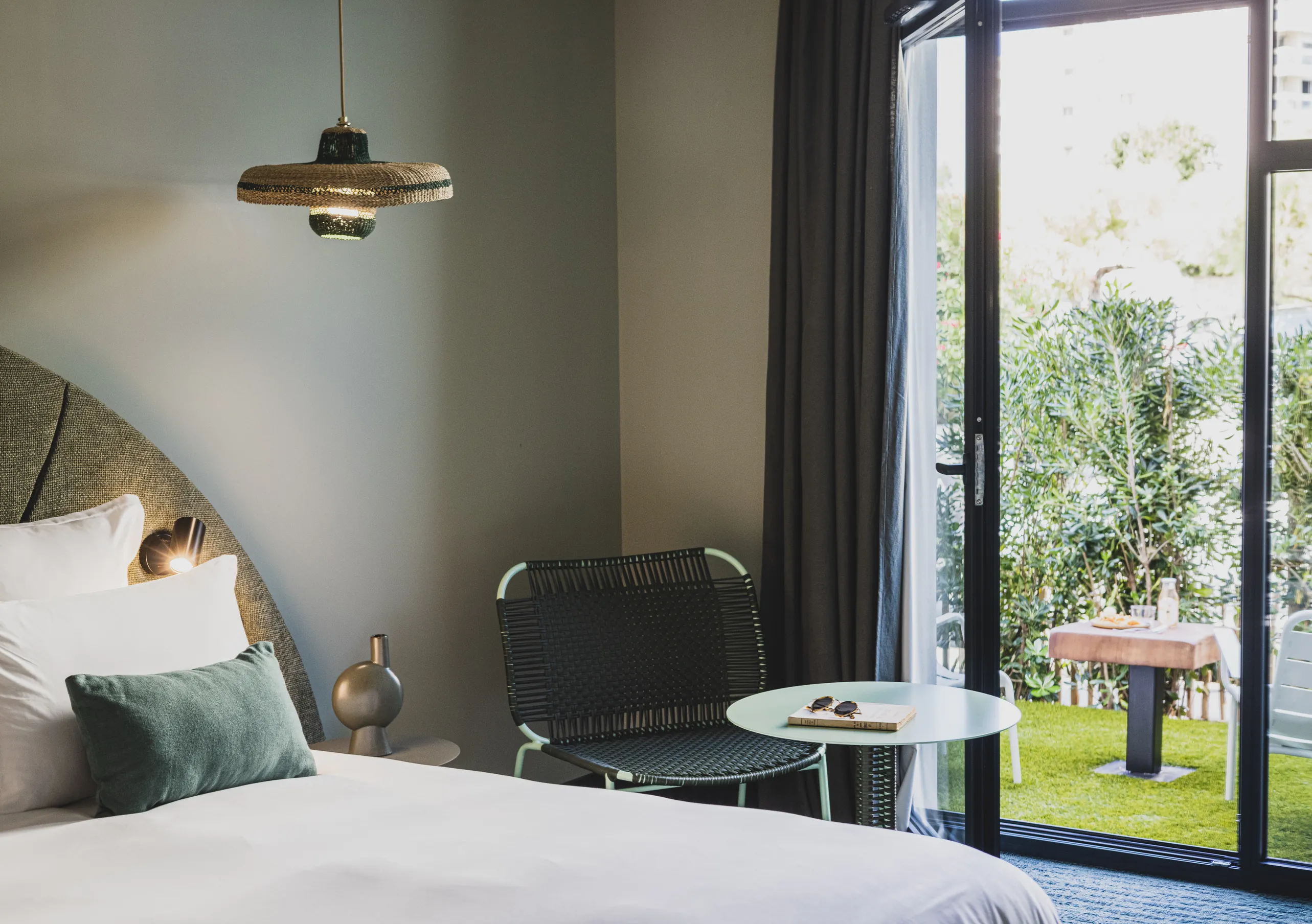 Le Garage Biarritz Experimental hotel room with olive green headboard, white bedding, industrial pendant lamp, and open glass door to private garden.