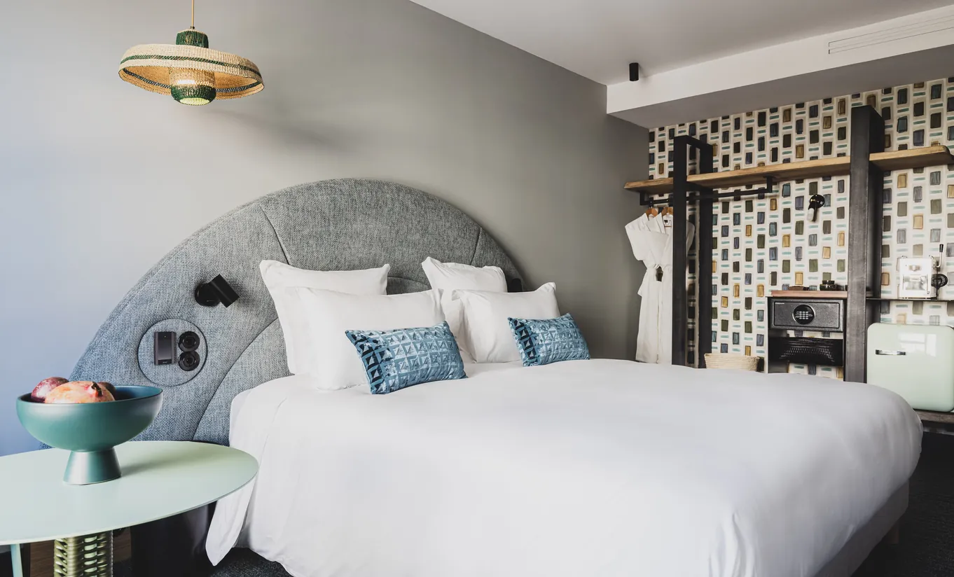 Le Garage Biarritz Experimental hotel suite with curved grey headboard, white bedding, blue cushions, decorative pendant light, and geometric room divider