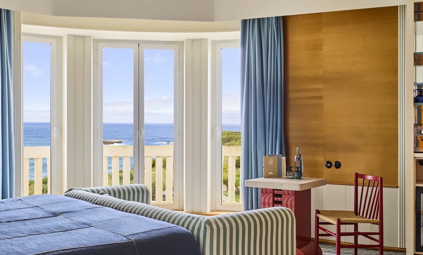 Regina Experimental Biarritz hotel sea-facing room with blue curtains, warm wood desk, two chairs, and full-width window with direct ocean panorama