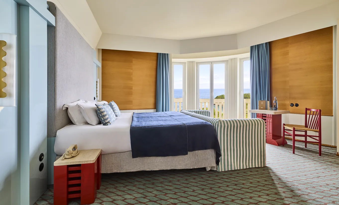 Regina Experimental Biarritz hotel room with warm wood wall paneling, blue bedspread, red side chair, and bright blue curtains