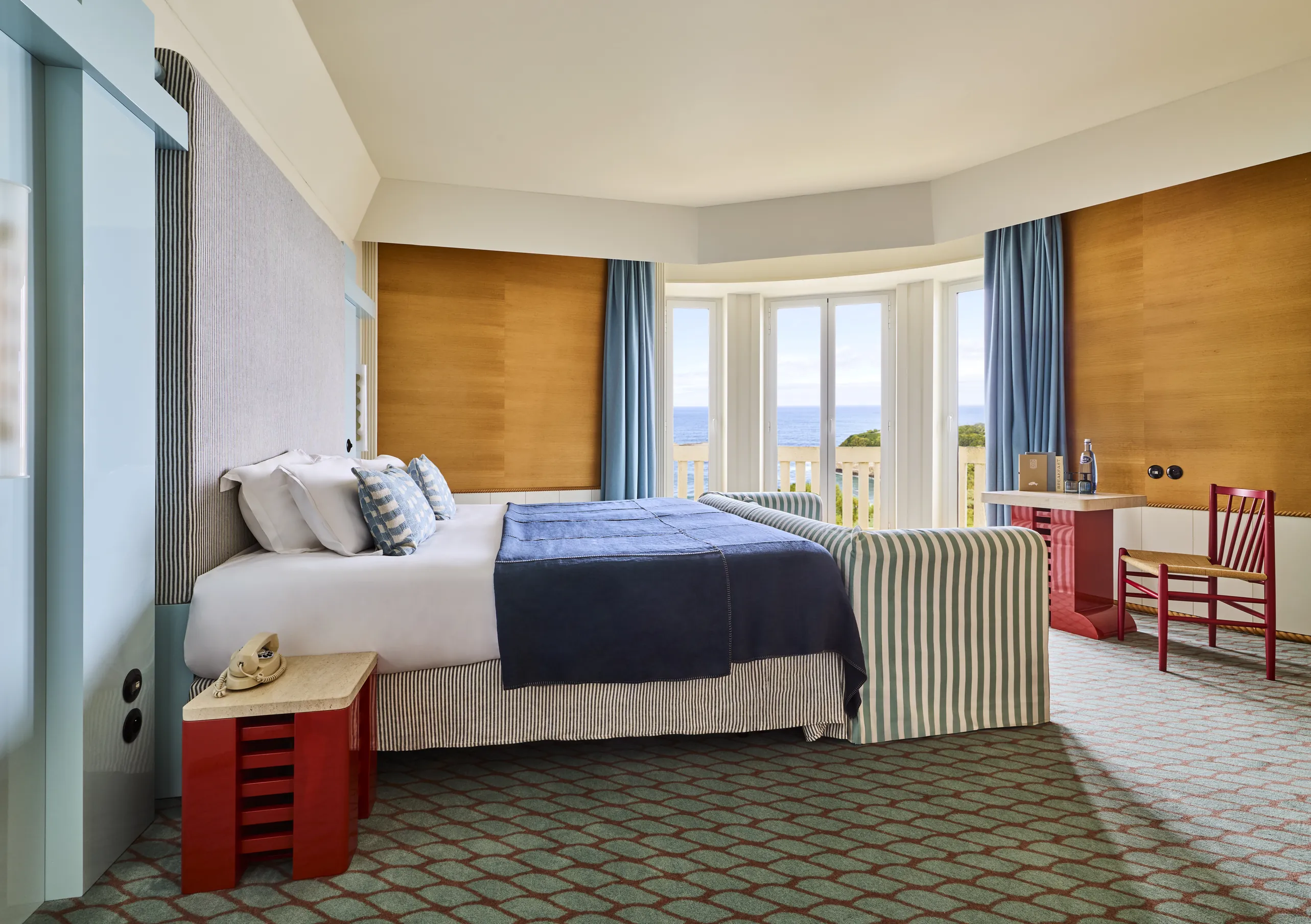Regina Experimental Biarritz hotel room with warm wood wall paneling, blue bedspread, red side chair, and bright blue curtains.