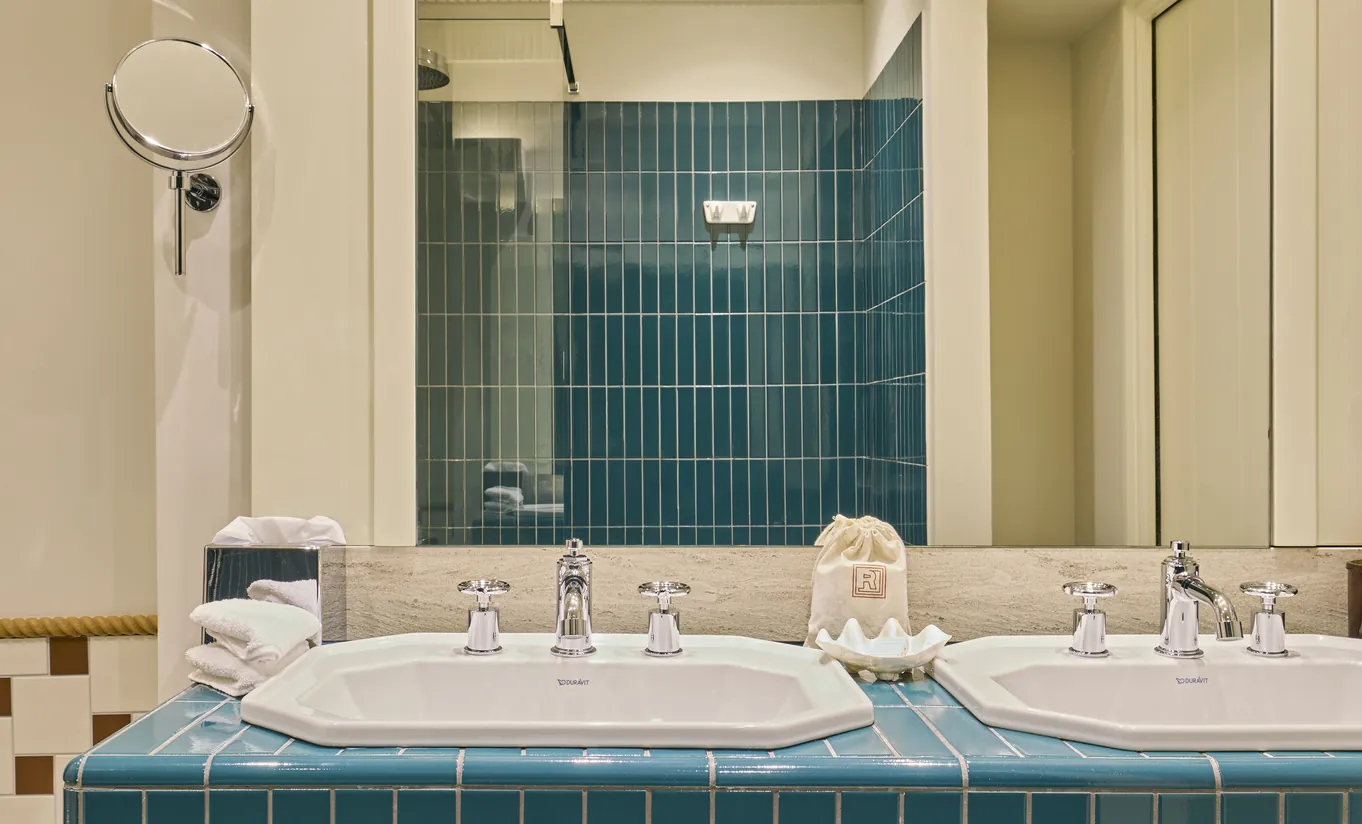 Experimental Group hotel bathroom with teal green mosaic tiled walls, double white sinks, and large vanity mirror with soft lighting